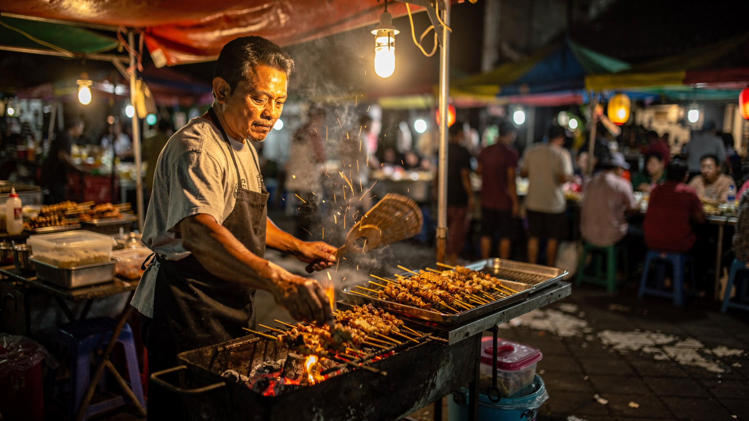 Endonezya'nın Yogyakarta şehrindeki hareketli bir pazarda, bir sokak yemekleri satıcısı kömür ızgarasında ustaca satay şişleri hazırlıyor, duman yükseliyor ve arka planda yerel müşteriler var.