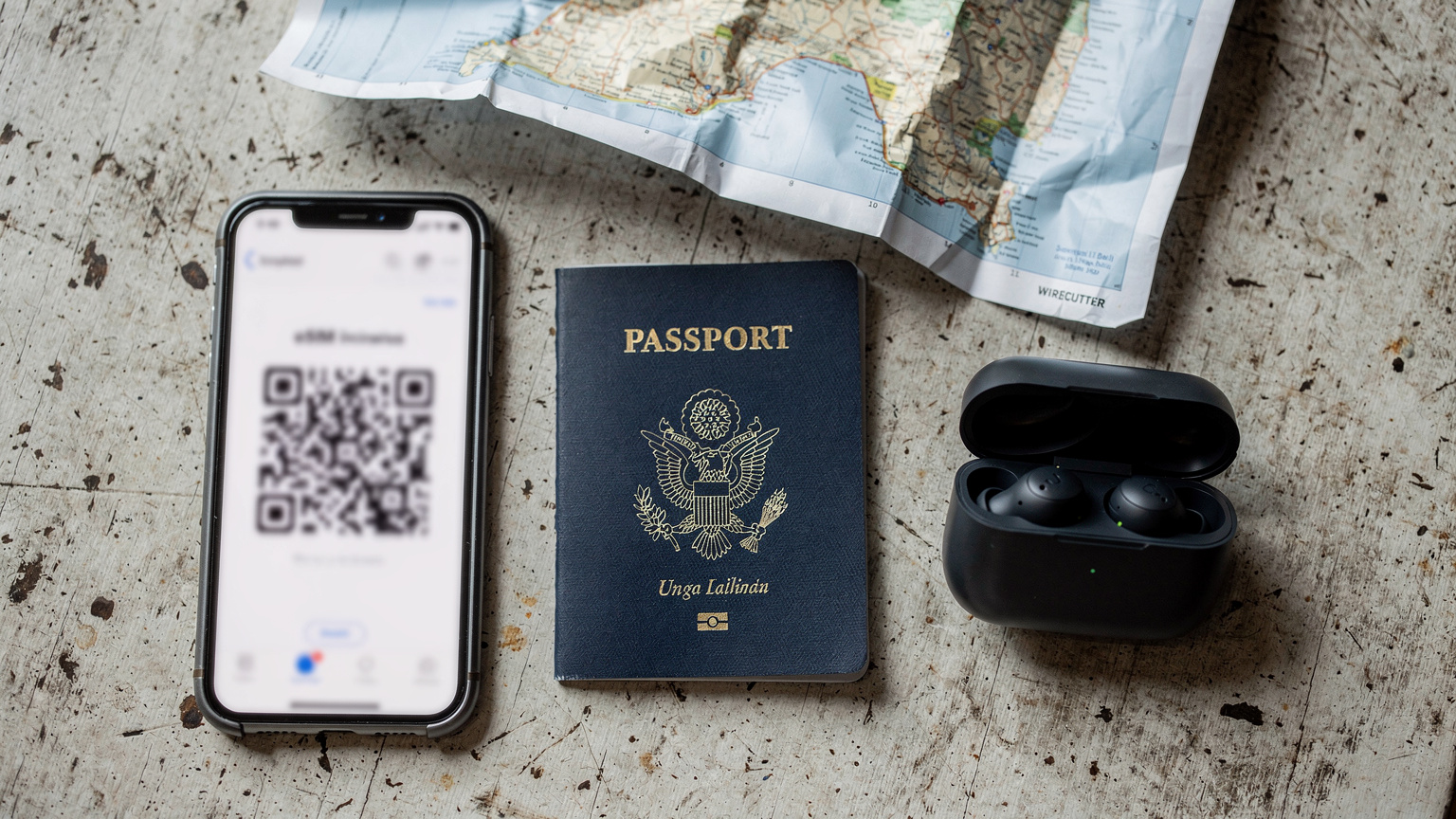 Pasaport, eSIM aktivasyonu için QR kodu gösteren bir akıllı telefon, bir çift kablosuz kulaklık ve buruşuk bir Bali haritası gibi temel seyahat eşyalarının tepeden çekilmiş bir fotoğrafı. Hepsi yıpranmış ahşap bir masanın üzerinde düzenli bir şekilde duruyor.