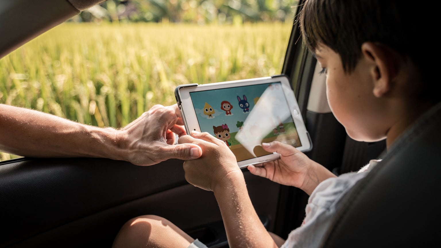 A close-up of a child's hands holding an iPad showing an offline game, with a parent's hand gently guiding them, inside a car in Bali.