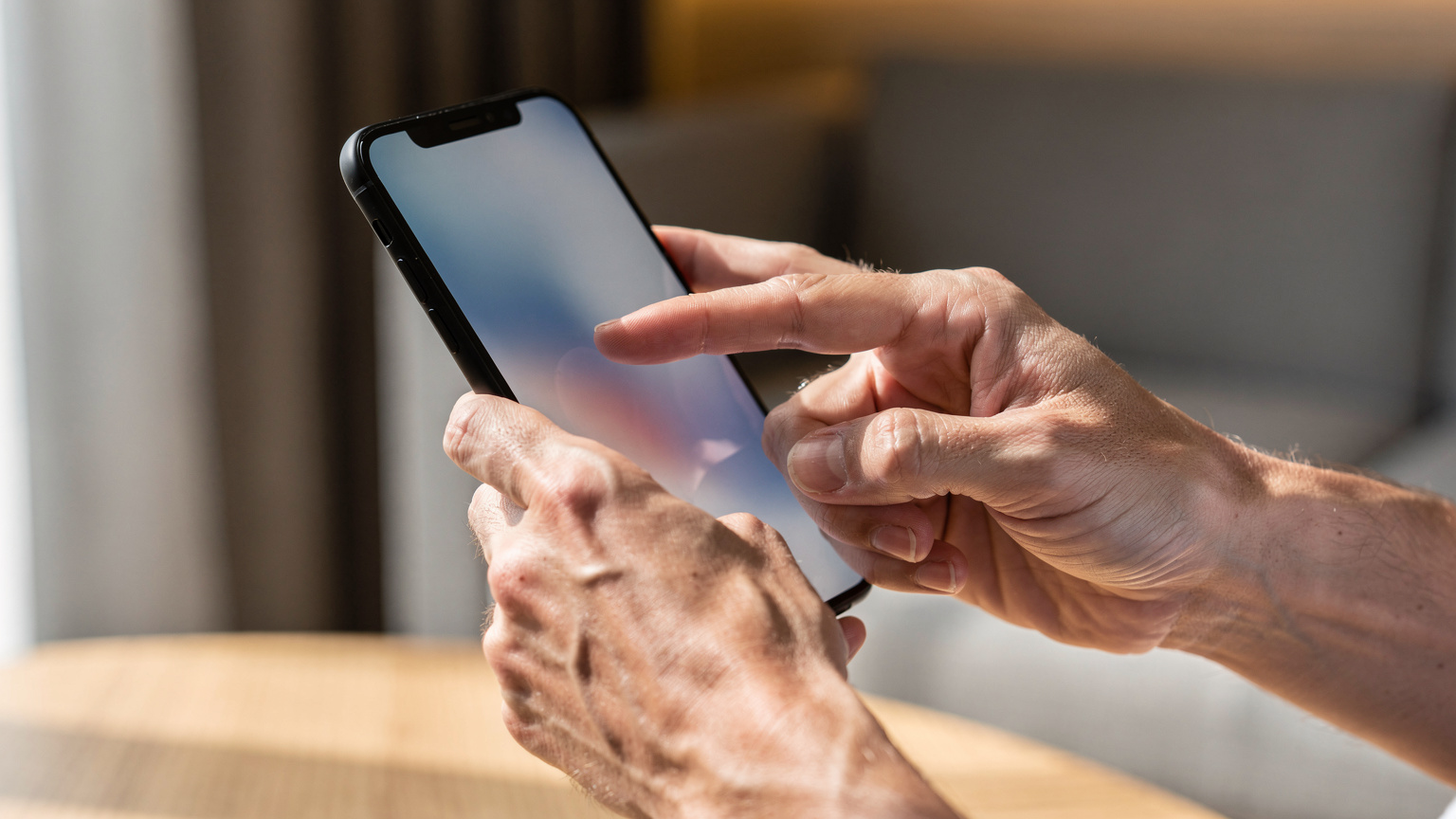 Close-up van twee handen die voorzichtig een smartphone vasthouden, waarbij de focus ligt op de gebaren van installatie of configuratie, met een wazige achtergrond van een modern interieur.