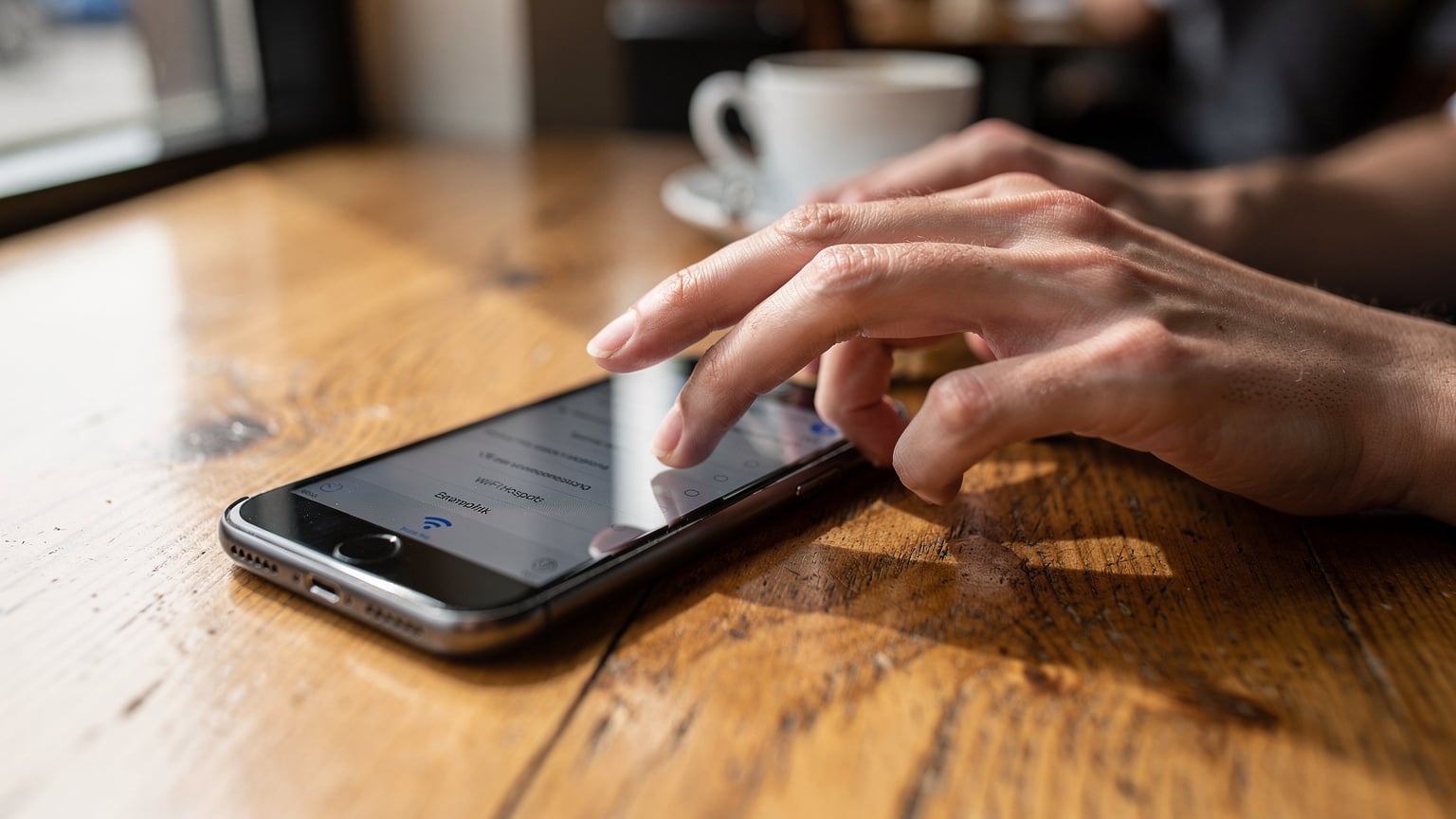 Bir kafenin ahşap masasında oturan bir kişinin elleri, akıllı telefonundaki Wi-Fi hotspot ayarları menüsünde gezinirken yakın çekim. Ekranı bulanık, sadece parmakların hareketi ve menü hiyerarşisi net.