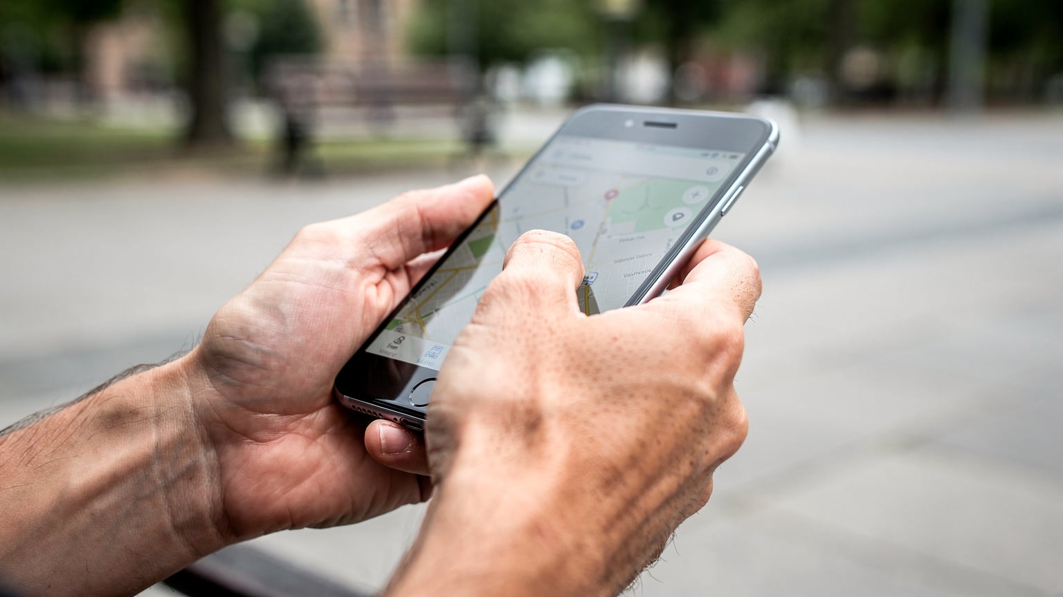 A close-up shot of a person's hands holding a smartphone, with their thumb scrolling through a map application, bright daylight, no UI text visible.