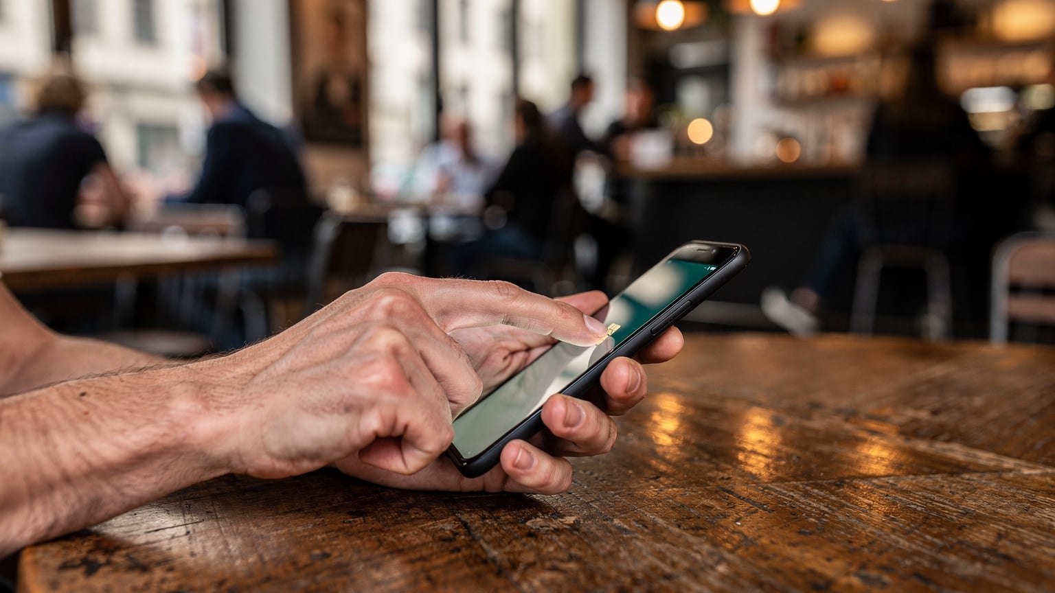 Nahaufnahme einer Hand, die ein Smartphone hält, während die andere Hand auf den Bildschirm tippt, um eine eSIM zu aktivieren. Die Person sitzt an einem Holztisch in einem belebten Café in Berlin-Mitte, mit unscharfem Hintergrund, der die Energie der Stadt andeutet.
