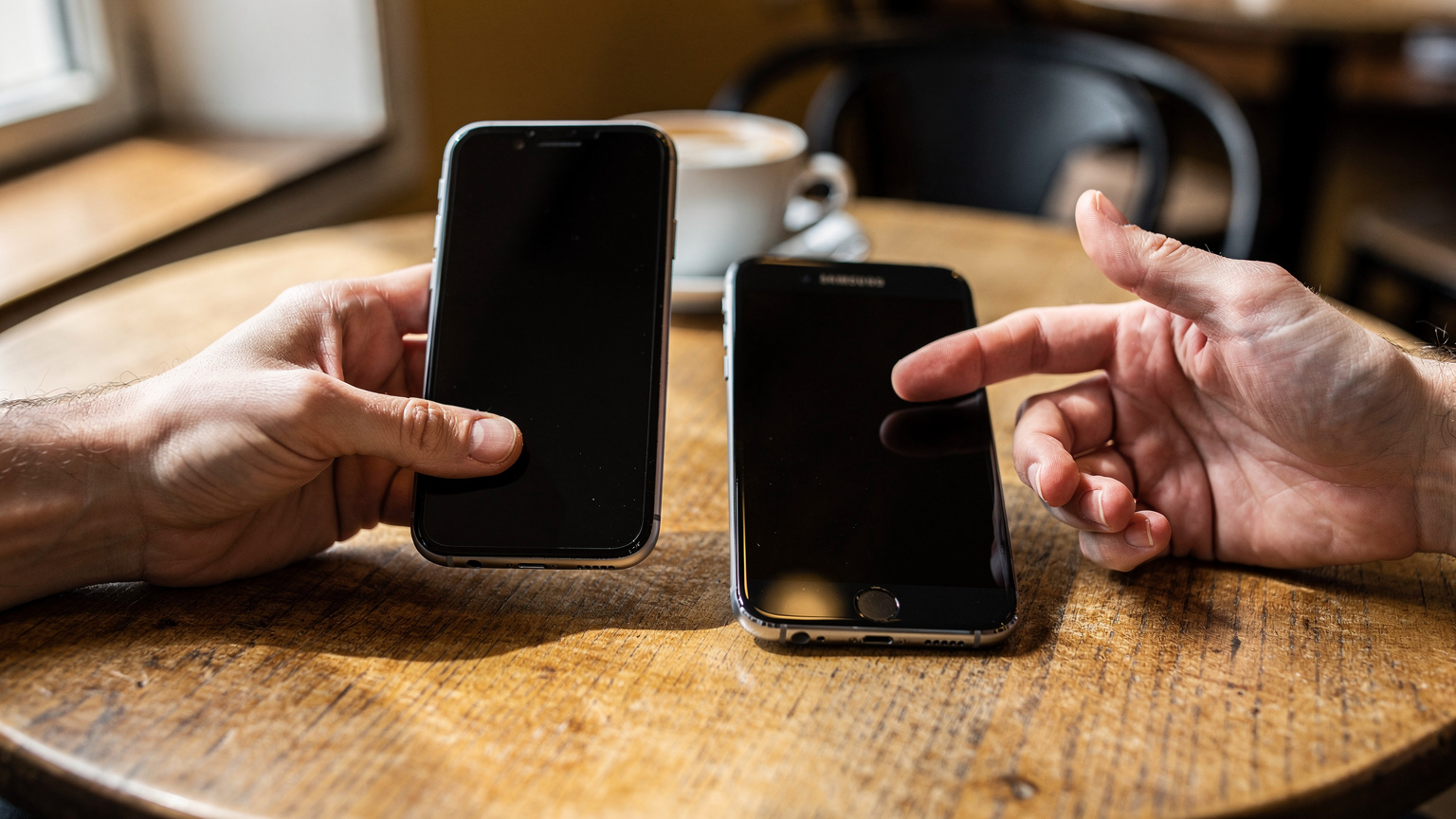 Händer som håller en iPhone och en Samsung sida vid sida på ett kafébord, tydligt att det pågår en enhetsbyte