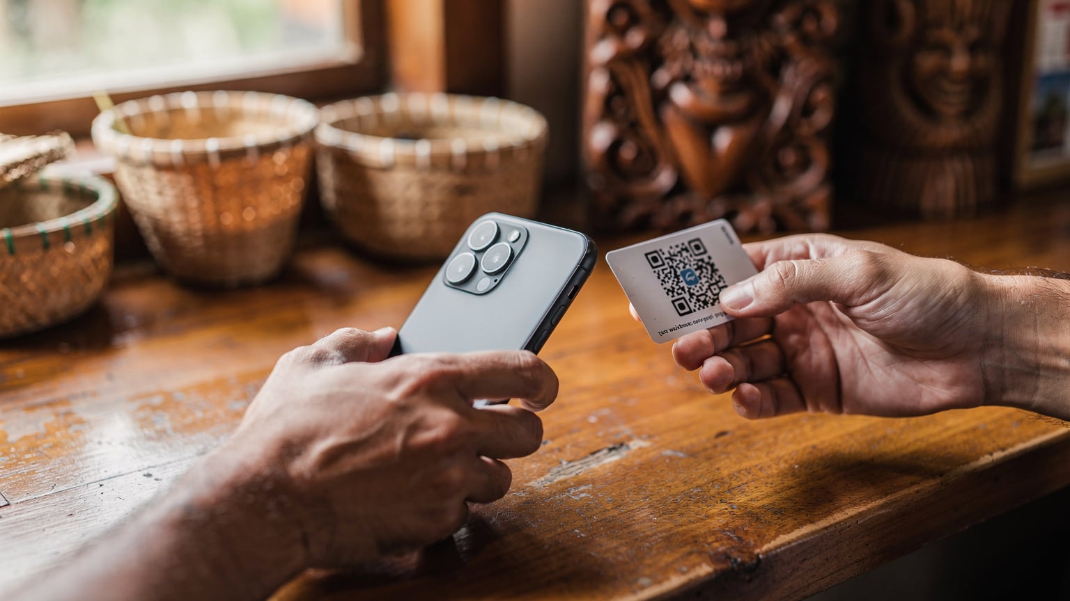 A close-up shot of hands using a smartphone to scan a QR code at a small, independent coffee shop counter in Ubud, Bali, with traditional Balinese woven baskets in the background.
