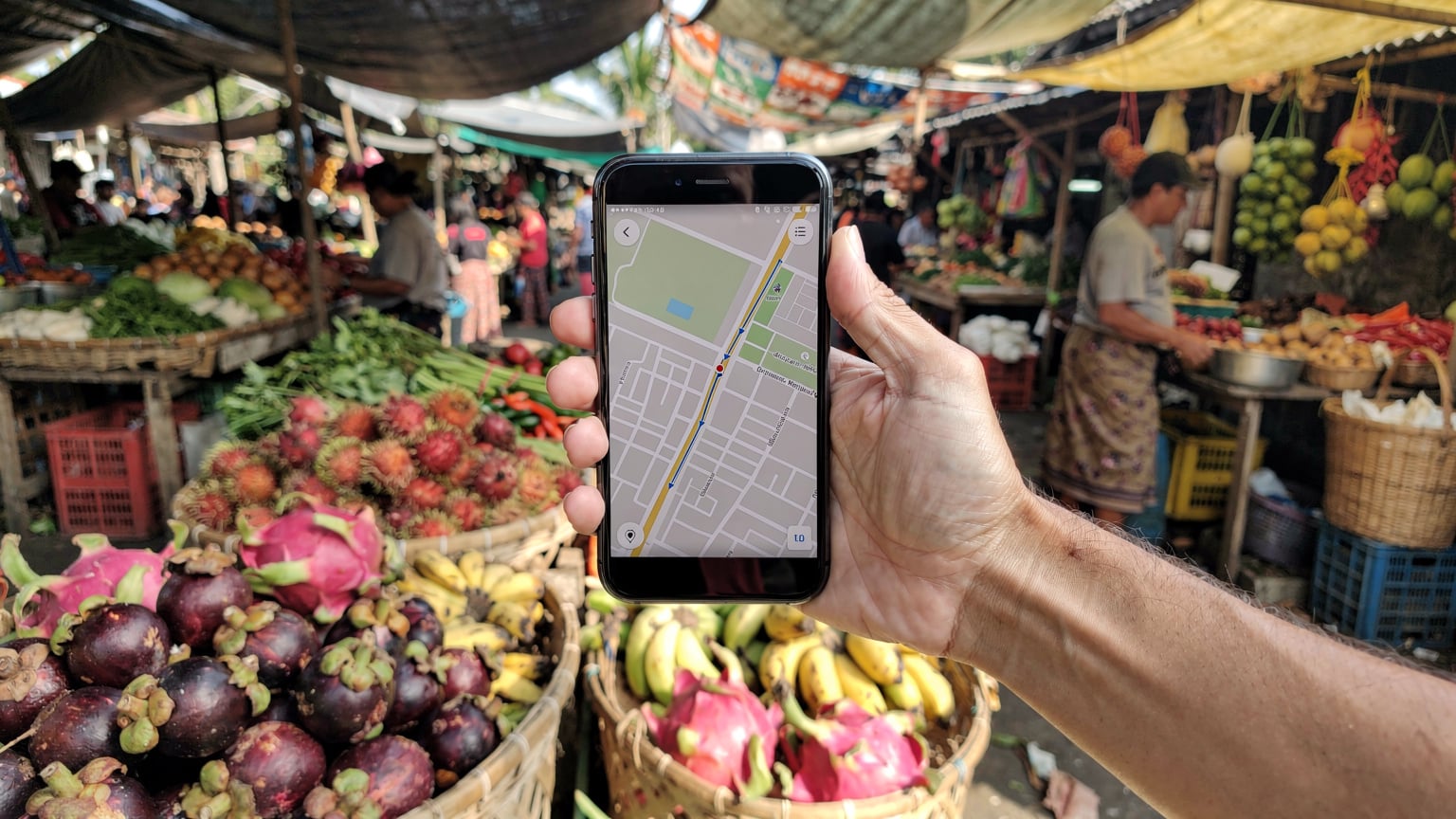 Bali'deki bir yerel pazar yerinde, taze meyve ve sebzelerin arasında, bir el akıllı telefonunu tutuyor, ekranında bir harita uygulaması açık.
