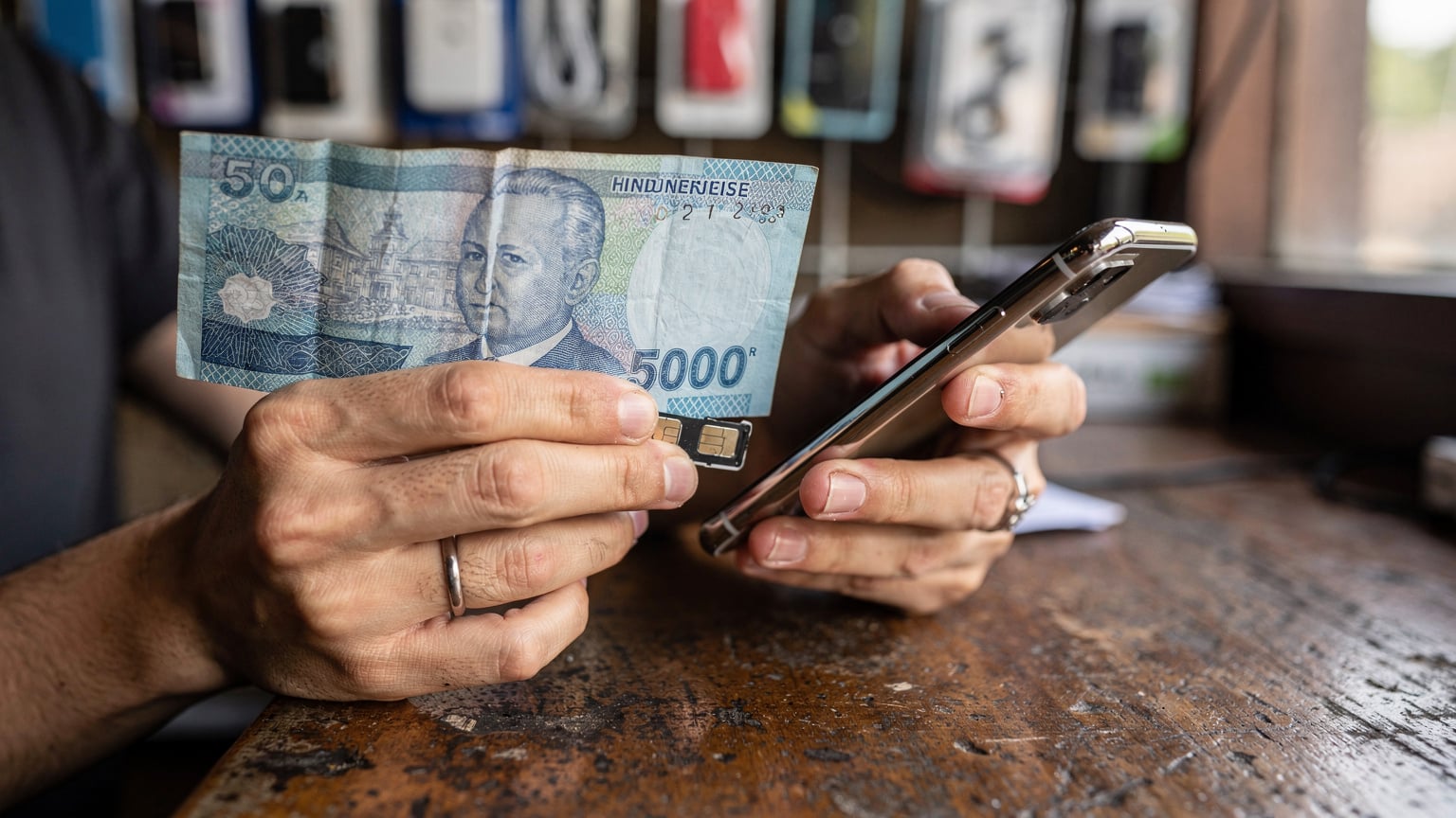 A close-up shot of a traveler's hands, one holding an Indonesian rupiah note and the other holding a smartphone with a SIM card tray open, preparing to insert a physical SIM card at a small, independent phone shop counter in Canggu, Bali.