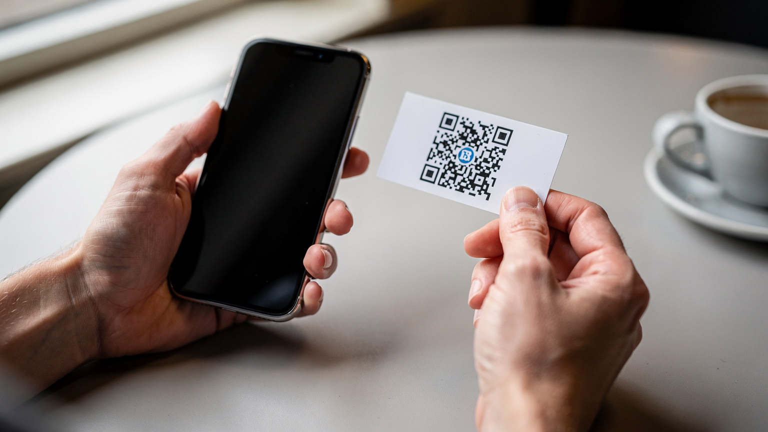 Een reiziger houdt een smartphone vast in de ene hand en scant met de camera een QR-code op een klein, wit papieren kaartje dat in de andere hand wordt gehouden. De focus ligt op de handen en de interactie.