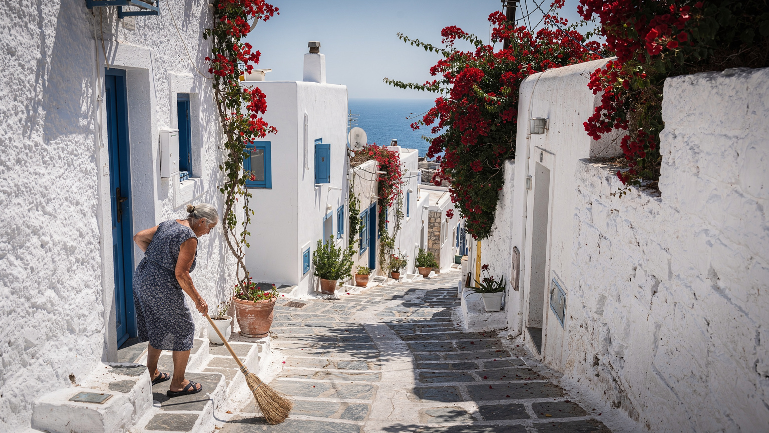 Στενό δρομάκι στη Χώρα της Μυκόνου με λευκά σπίτια, κόκκινες μπουκαμβίλιες και μια ντόπια κυρία να σκουπίζει το κατώφλι της το μεσημέρι.