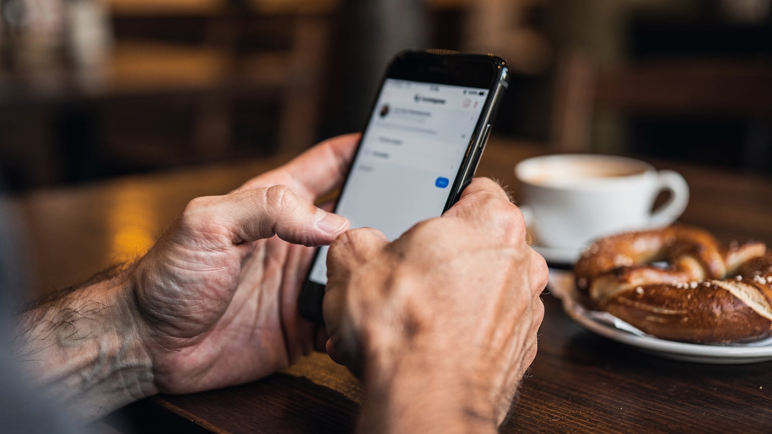 Nahaufnahme der Hände eines Mannes, der ein Smartphone hält und mit dem Daumen über den Bildschirm streicht, während er in einem Café in einem Münchner Stadtteil sitzt. Die Tischplatte ist aus dunklem Holz, im Hintergrund verschwommen eine Kaffeetasse und eine Brezel.