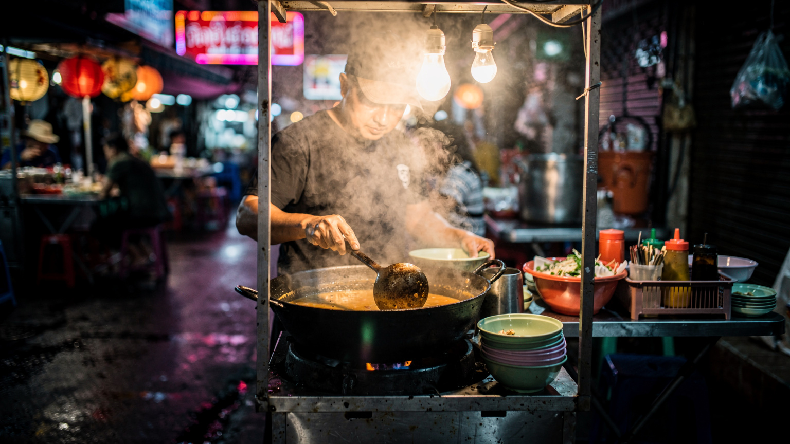 Penjual street food di kaki lima Bangkok menghidang sup tom yam panas, wap mengepul, suasana malam yang sibuk
