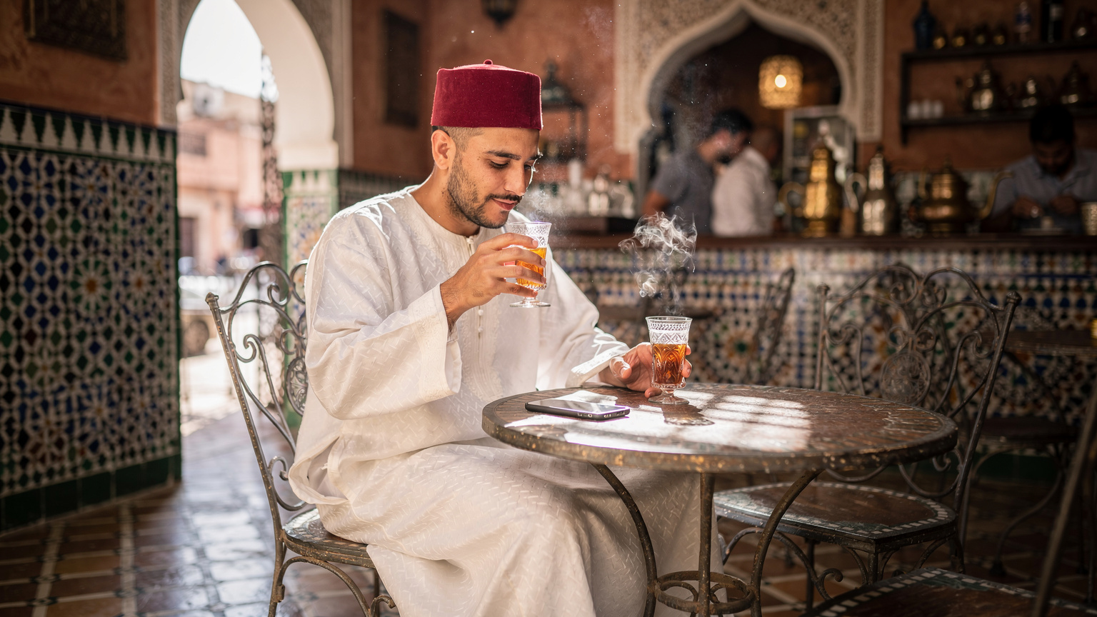 Un homme sirotant un thé à la menthe dans un café marocain traditionnel, utilisant discrètement son téléphone, avec des jeux de lumière matinale sur le sol en zellige.