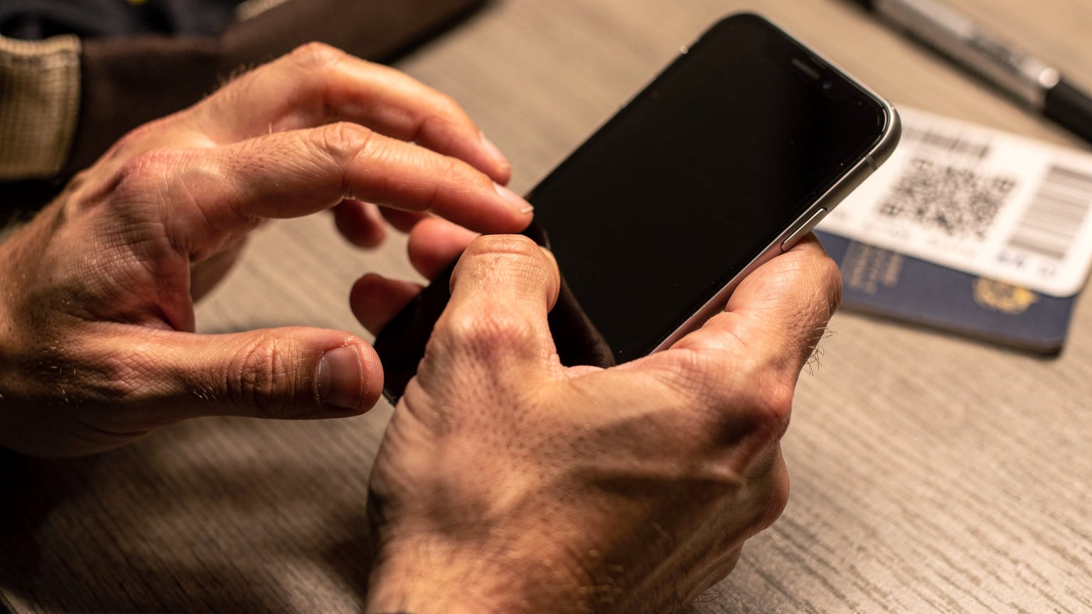 Nahaufnahme von Händen, die ein Smartphone halten und einen QR-Code scannen, mit einem unscharfen Hintergrund von Reisepässen und einer Bordkarte auf einem Tisch.