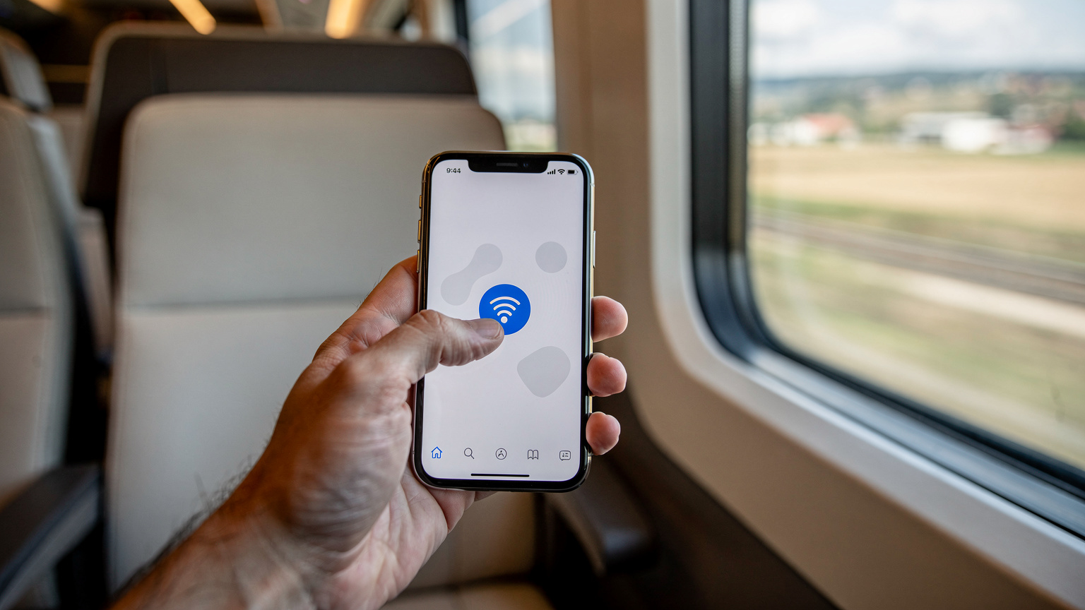 Bir tren kompartımanında, bir el iPhone ekranında Wi-Fi Hotspot ayarlarını açıyor, arka planda tren penceresinden geçen manzara flu.