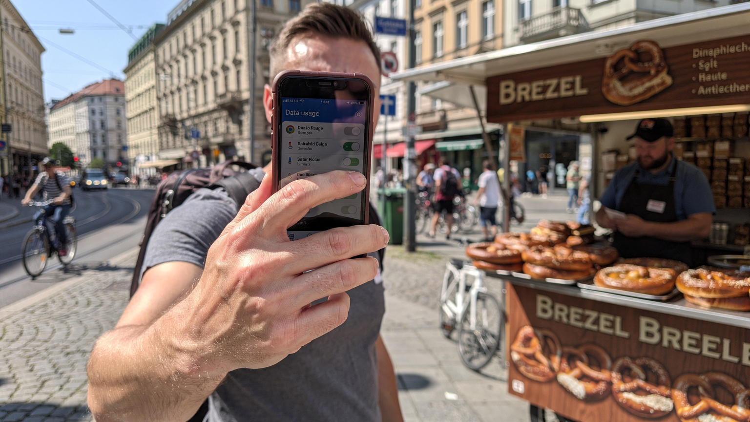 Berlin'de hareketli bir sokakta, bir el telefonda bir eSIM sağlayıcısının uygulama ekranında veri kullanımını kontrol ediyor, arka planda bir sokak satıcısı Brezel satıyor.
