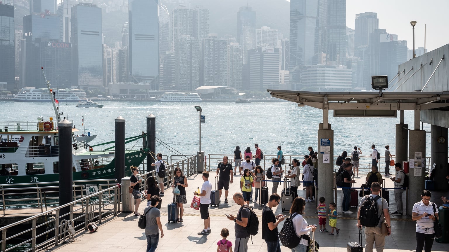 香港繁忙的尖沙咀码头，旅客们正在等待渡轮，背景是维多利亚港和香港岛的摩天大楼。码头人潮涌动，展现出城市的活力。