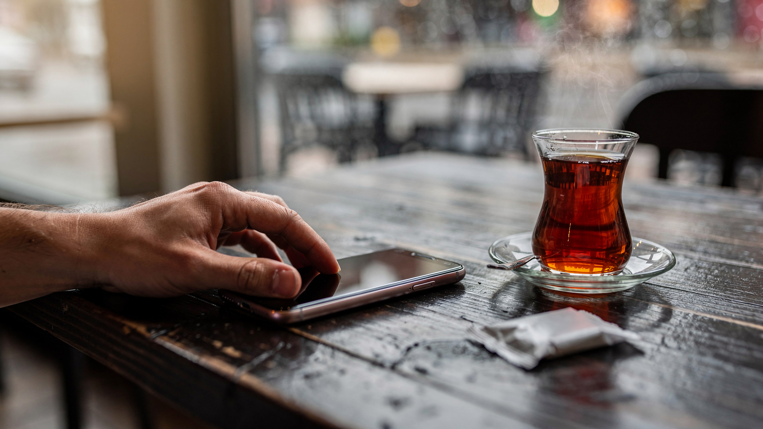 Mano che appoggia un telefono a schermo spento su un tavolo di legno in un caffè, con un tè turco vicino