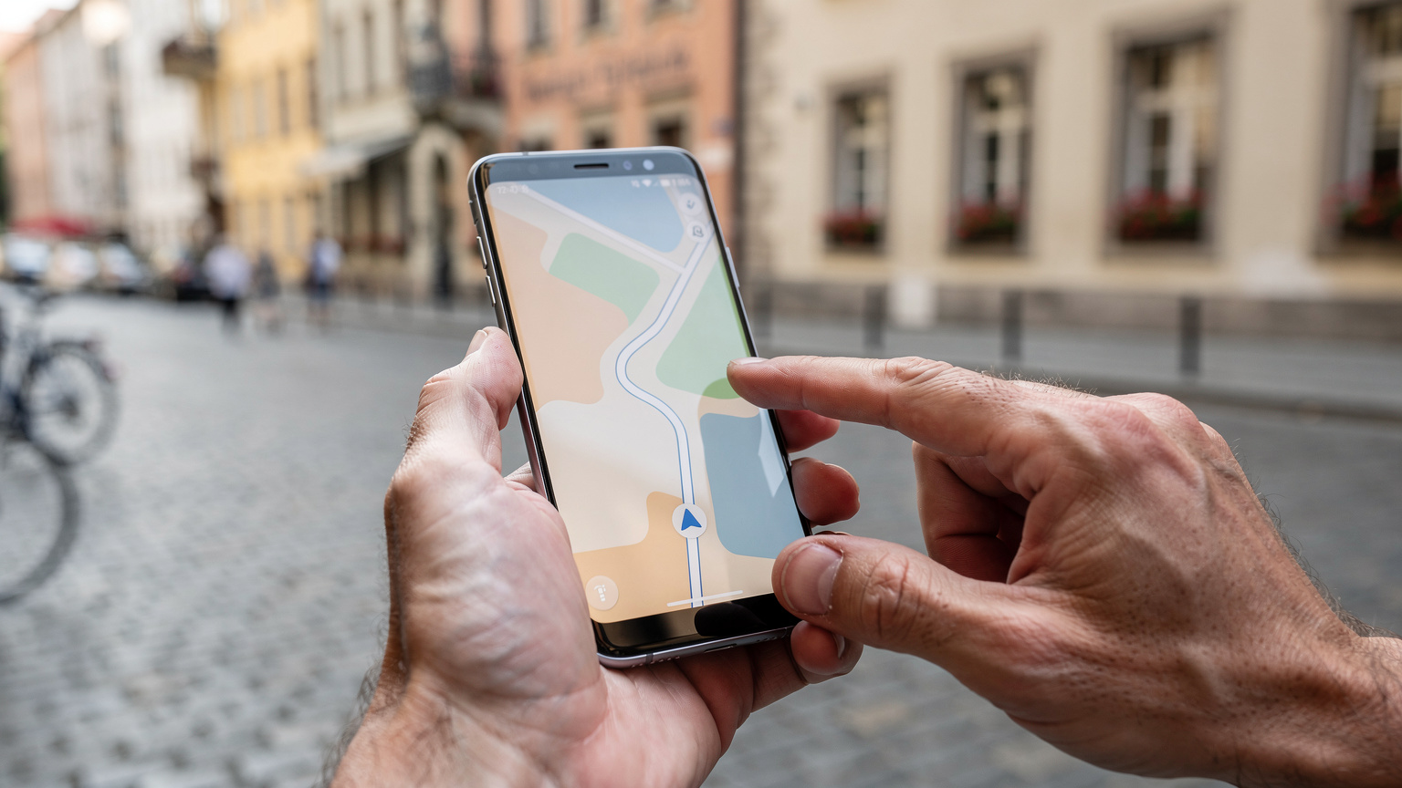 Zwei Hände halten ein Smartphone, auf dessen Bildschirm eine detaillierte Straßenkarte mit einer geplanten Reiseroute angezeigt wird. Im unscharfen Hintergrund ist eine europäische Stadt zu erkennen.