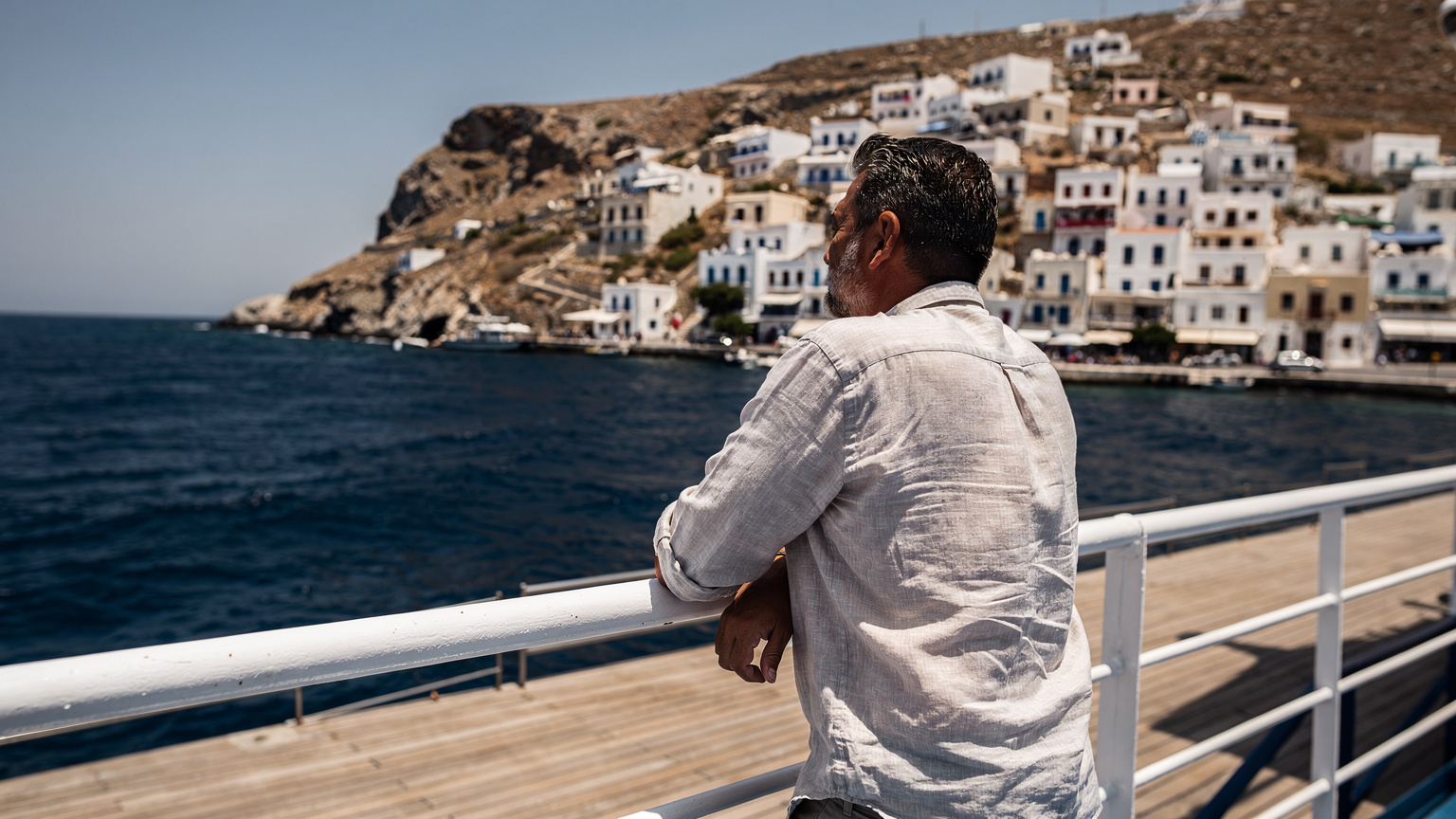 Επιβάτης στο κατάστρωμα του Blue Star κοιτάει τη θάλασσα, με την Τήνο στο βάθος, μεσημεριανό φως με σκληρές σκιές.