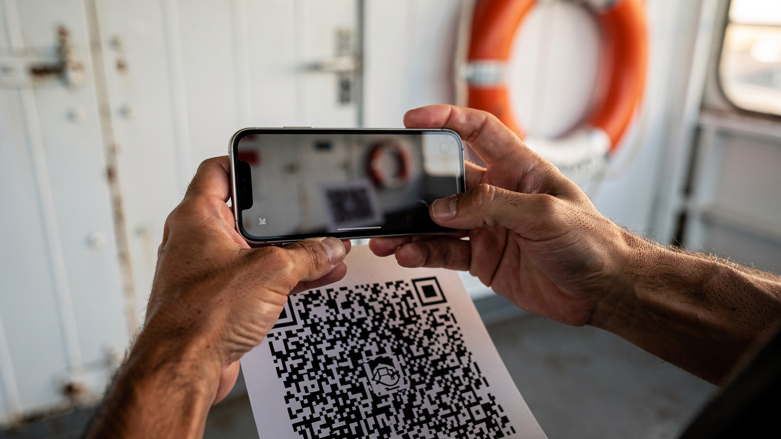Κοντινό πλάνο χεριών που κρατούν iPhone πάνω από εκτυπωμένο QR code, με φόντο το κατάστρωμα πλοίου.