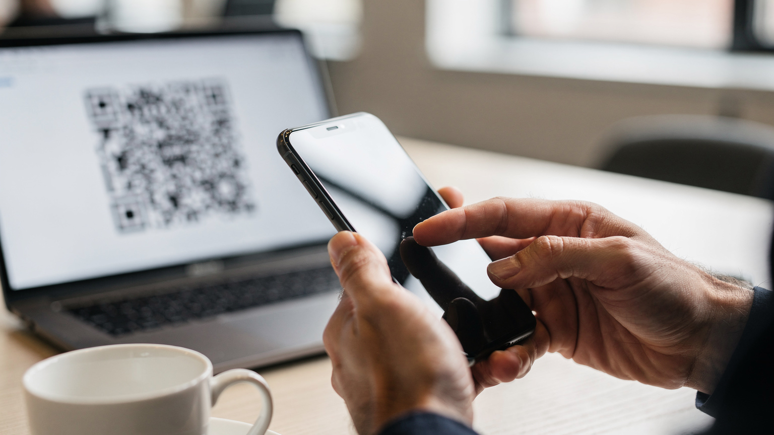 Primo piano di una mano che tiene uno smartphone, mentre l'altra scorre sullo schermo per installare una nuova eSIM, con un codice QR leggermente fuori fuoco sullo sfondo su un monitor di laptop in un coworking.