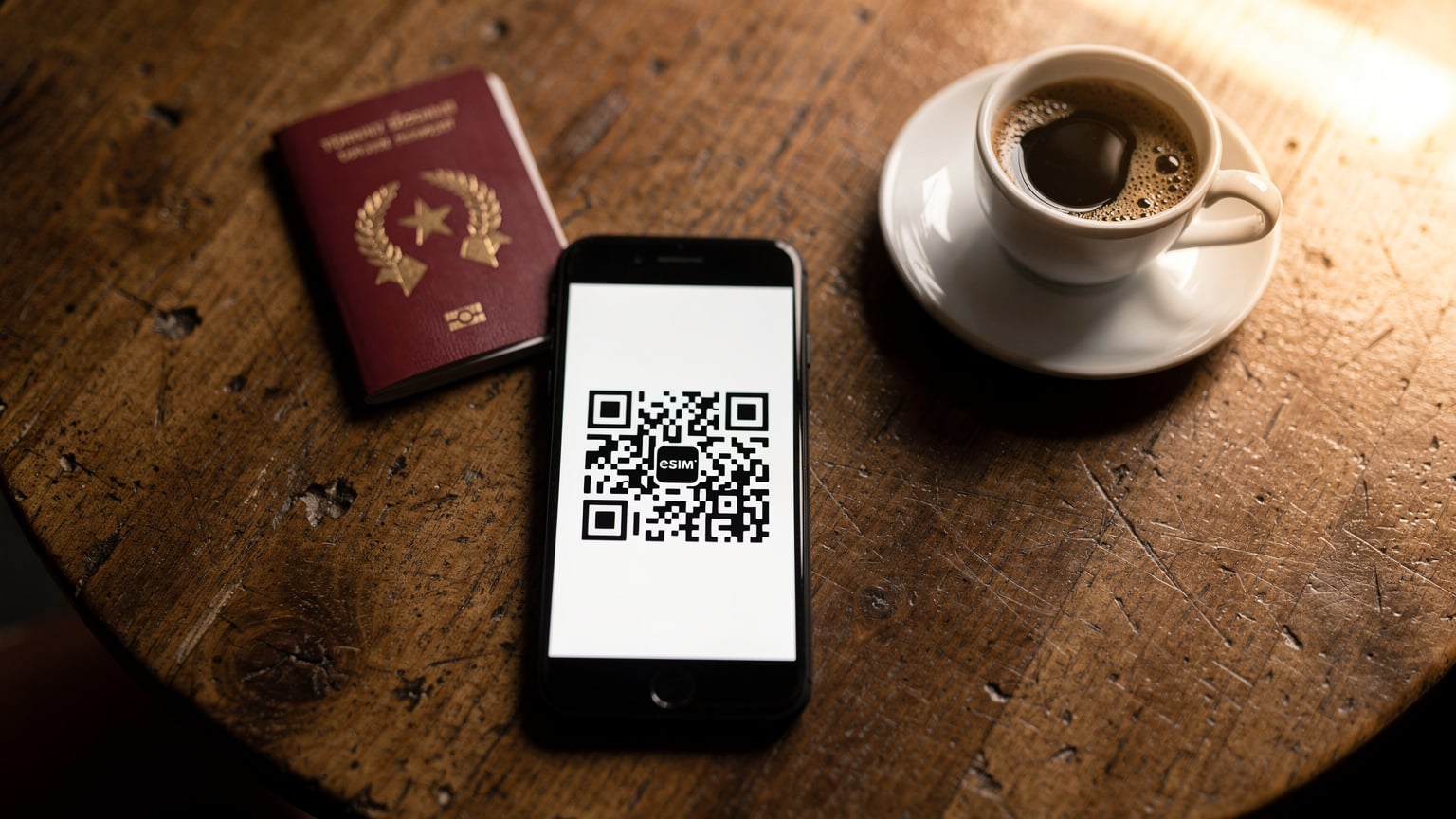 Bir kafenin ahşap masasında duran akıllı telefon ekranında bir eSIM kurulum QR kodu, arka planda bulanık pasaport ve kahve fincanı olan bir flat-lay fotoğrafı.