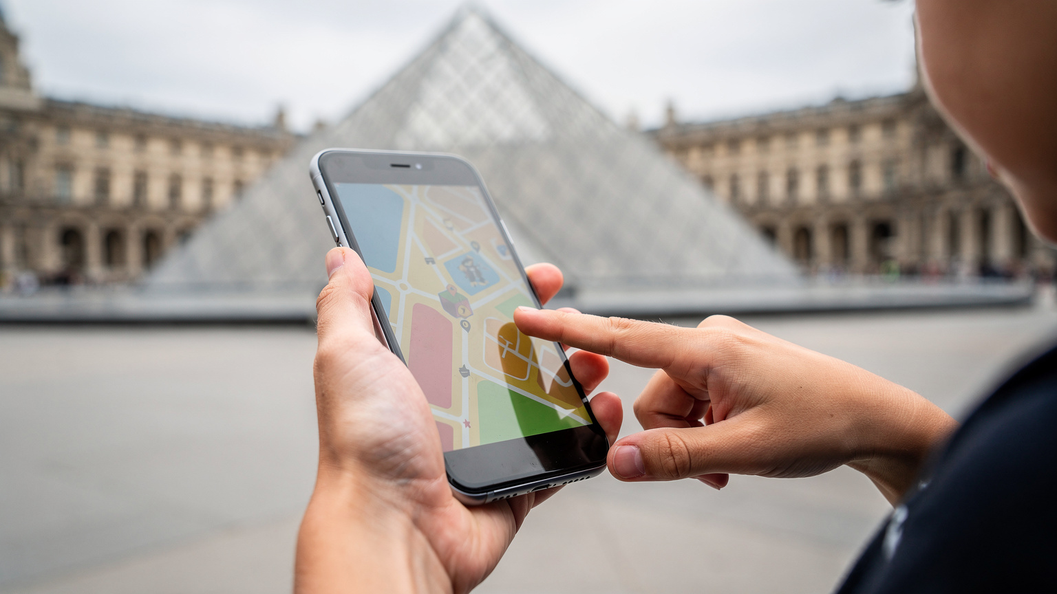 Paris'teki Louvre Müzesi'nin avlusunda, bir annenin ve çocuğunun elleri, açık bir harita uygulamasını gösteren bir akıllı telefonu inceliyor, arka planda müzenin piramidi hafifçe seçiliyor.