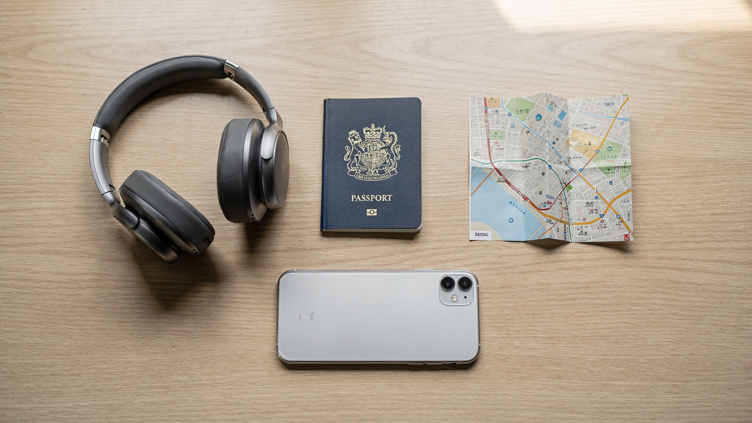 A close-up, editorial flat-lay of a neatly organized travel kit: a passport from the UK, a pair of wireless headphones, a small folded paper map of Seoul, and a smartphone lying face-down, all on a clean wooden table.
