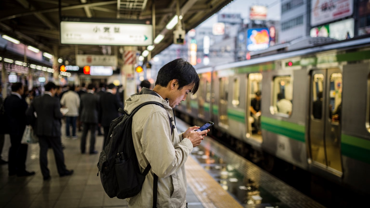 Un viaggiatore consulta il suo telefono mentre aspetta il treno sulla banchina affollata della stazione di Shibuya a Tokyo, con la frenesia della città sullo sfondo e i neon luminosi che si riflettono sul binario bagnato.