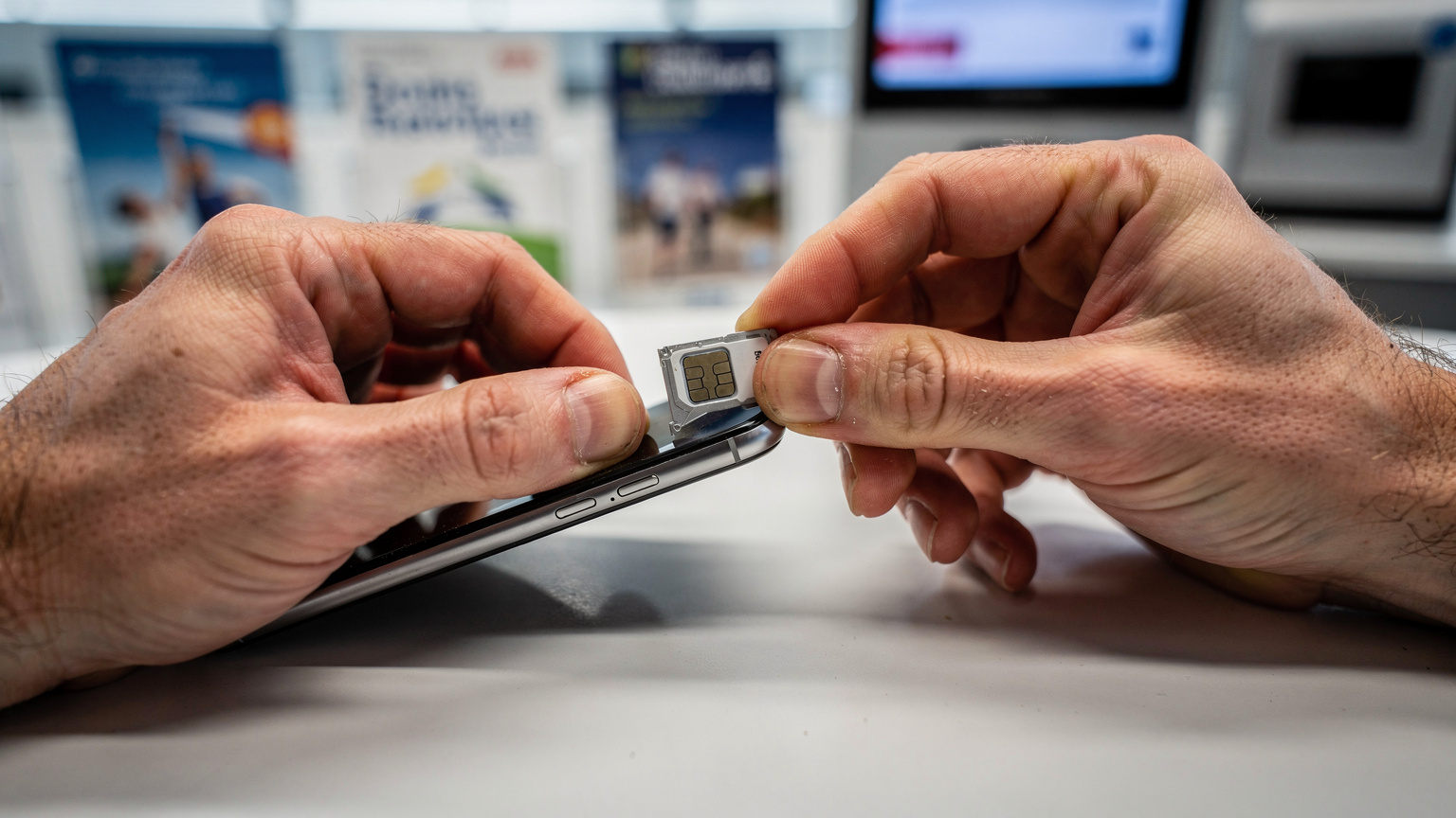 A close-up shot of a pair of hands struggling to insert a tiny physical SIM card into a phone's SIM tray at a brightly lit airport kiosk, with frustrated body language.