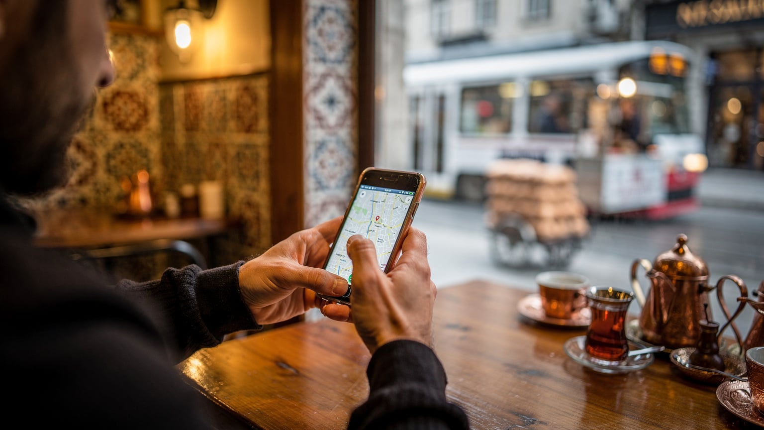 Bir kafenin penceresinden görünen hareketli İstanbul sokağında, bir kişi elinde akıllı telefonuyla yol tarifi bakıyor. Telefon ekranı görünmüyor, sadece kullanım anı vurgulanıyor.