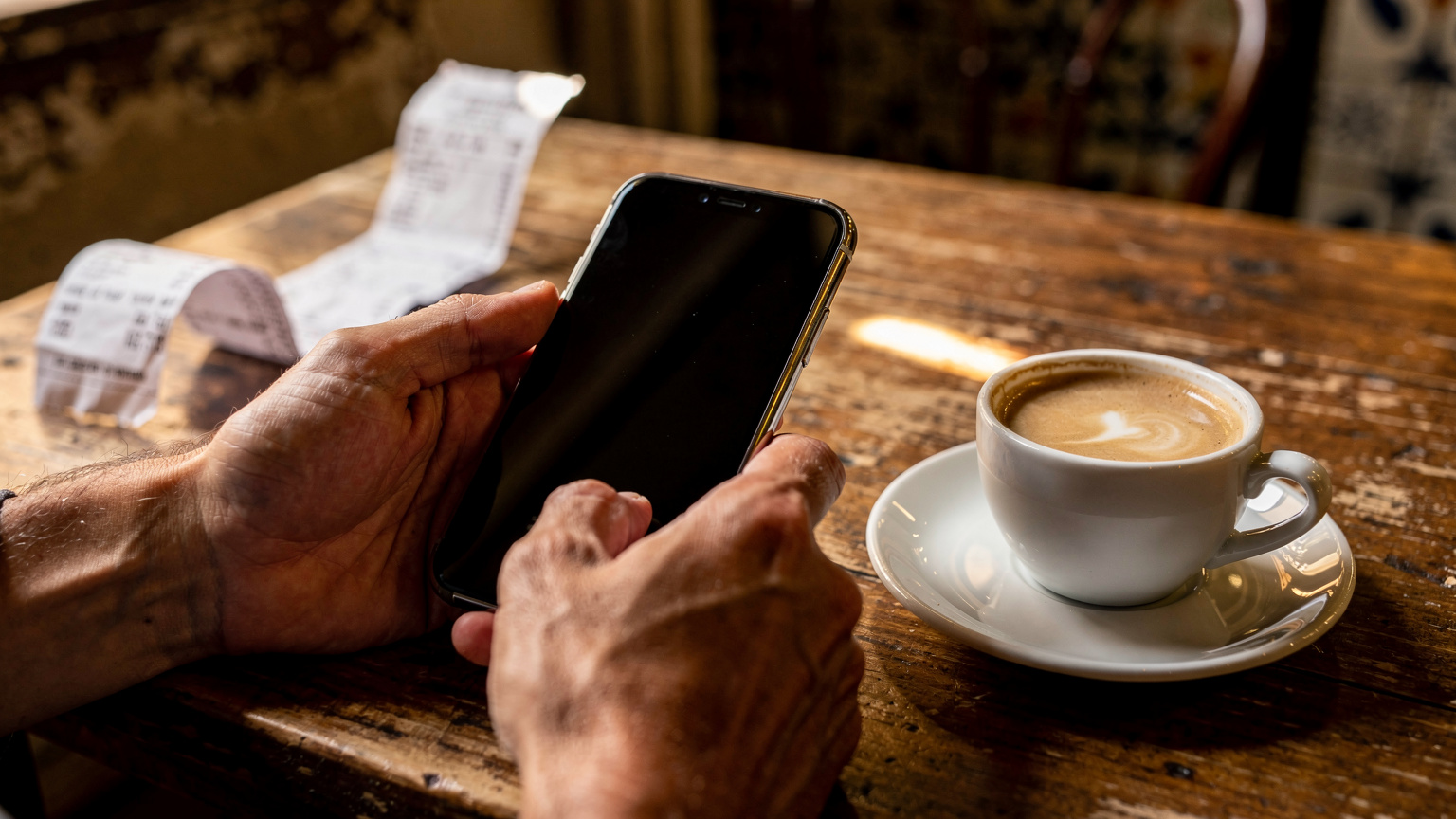 Manos de viajero sosteniendo un teléfono apagado junto a un café con leche en un bar de Asunción
