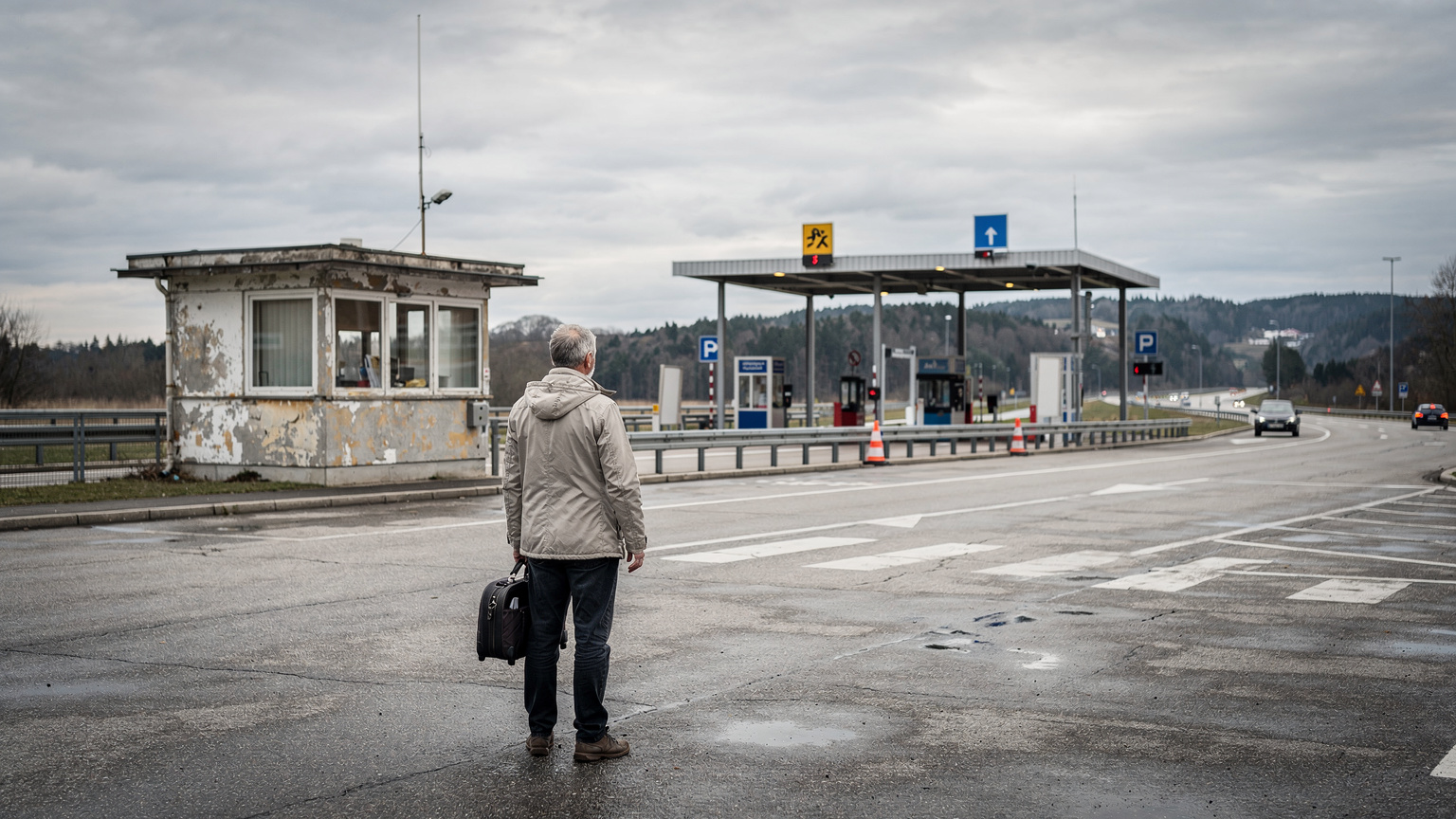 Ein Reisender steht an einem Grenzübergang zwischen Deutschland und Österreich auf einem leeren Parkplatz, im Hintergrund ist ein kleines, verlassenes Zollhäuschen und die Autobahn zu sehen.