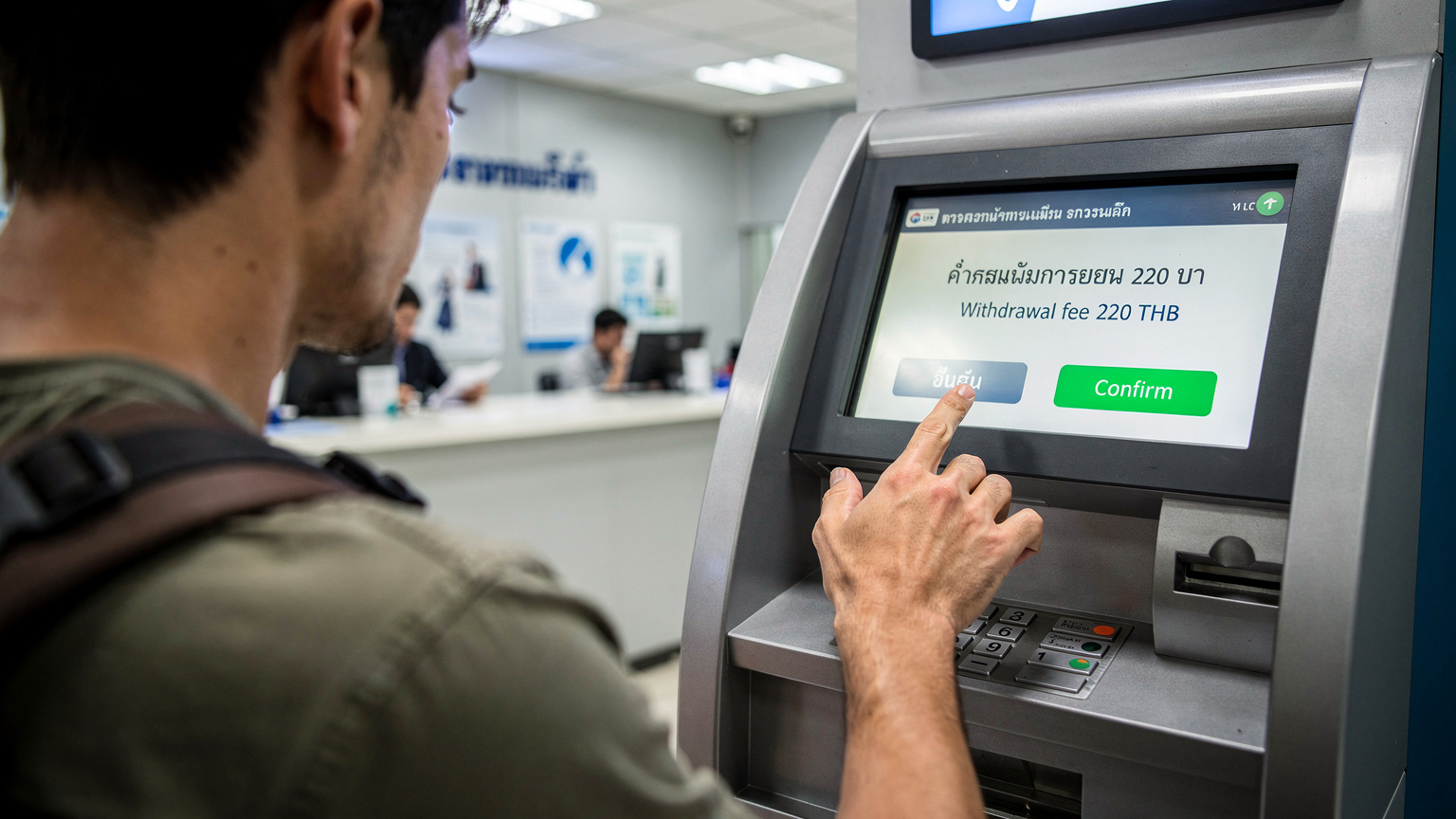 En resenär granskar noggrant en bankomatskärm i en bank i Bangkok, där avgifterna för uttag visas tydligt på thai och engelska, i starkt lysrörsljus från taket.