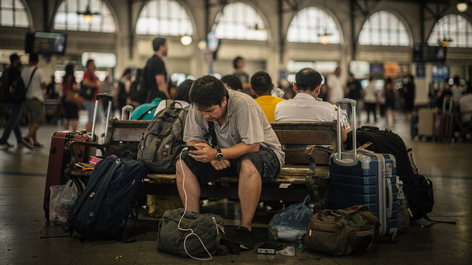 En person försöker ladda sin telefon med en power bank på en överfull bänk på en tågstation i Bangkok, omgiven av bagage och andra resenärer i ett kaotiskt men typiskt transithallsscenario.