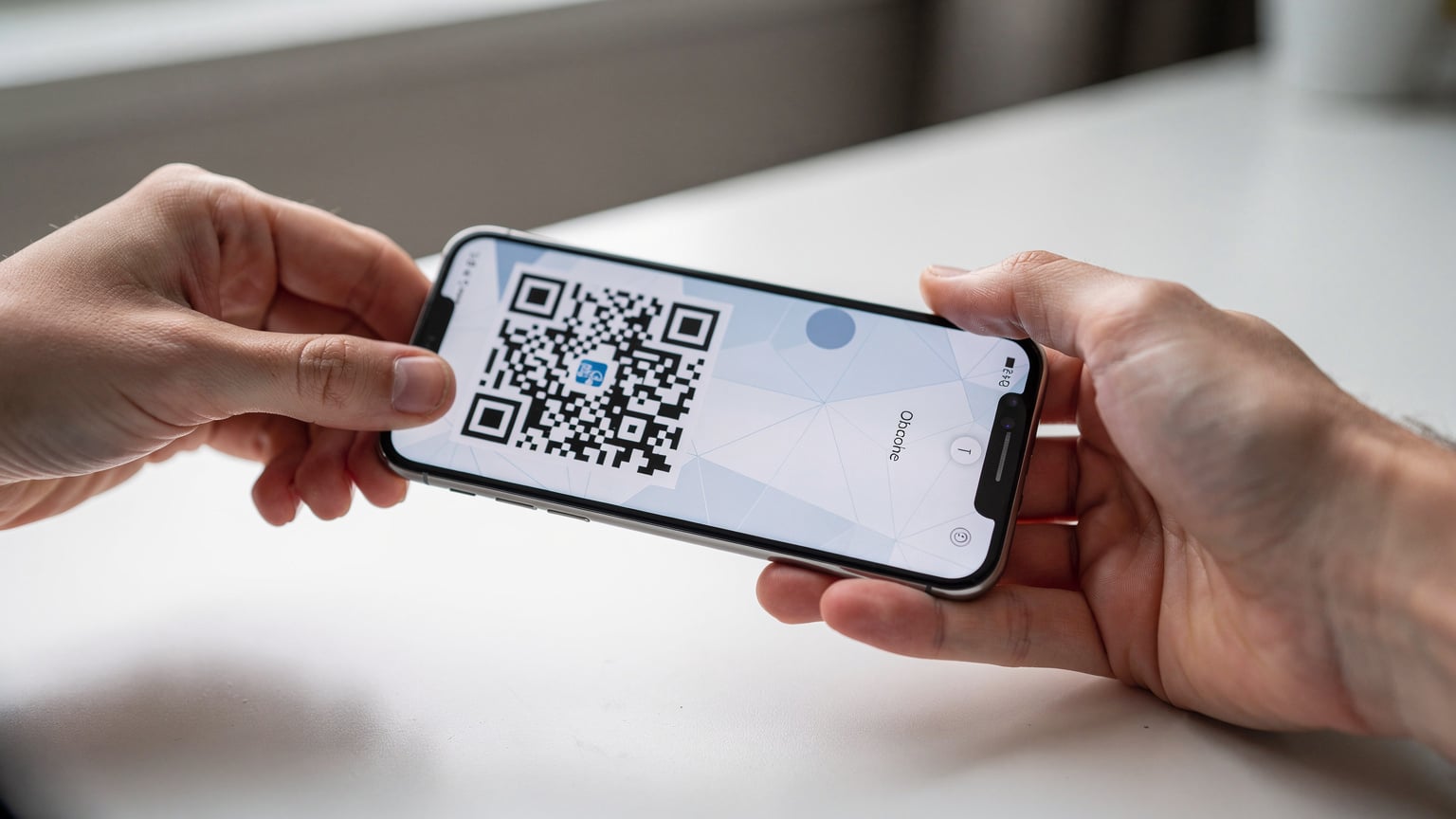 Bir akıllı telefon ekranında Türk Telekom eSIM kurulumu için QR kodu tarama arayüzü gösterilirken, bir el kodu cihaza doğru tutuyor. Ekranda okuma güçlüğü çekilen soyut geometrik desenler var.