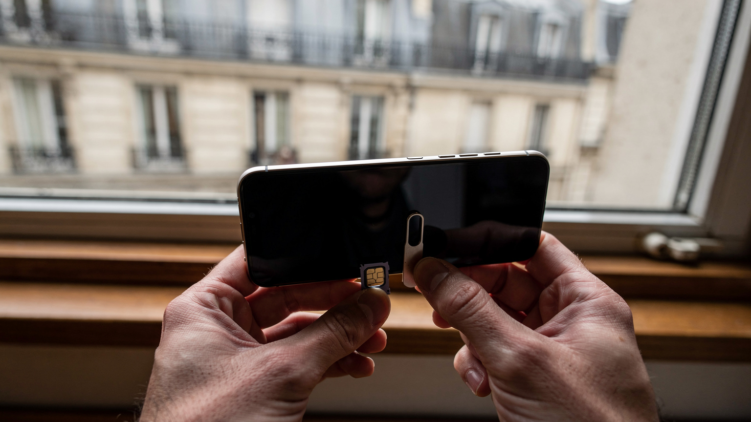Close-up de mãos a segurar um telemóvel com uma ferramenta de extração de SIM a abrir a bandeja, no parapeito de uma janela em Paris.