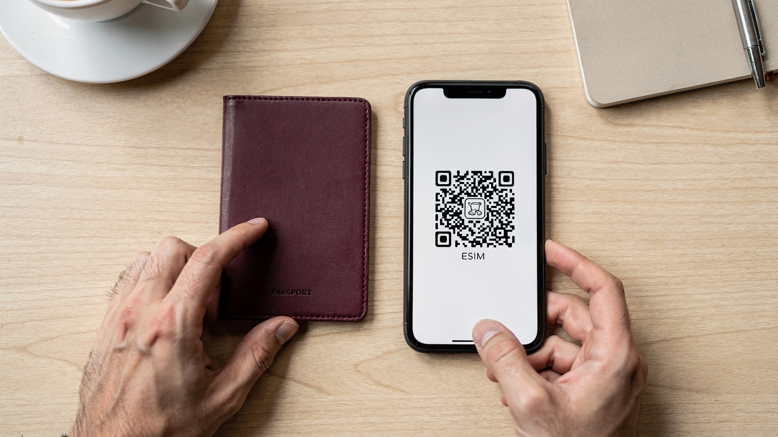 Bir dijital göçebe, pasaportunu ve üzerinde bir eSIM QR kodu olan akıllı telefonunu masanın üzerinde gösteriyor. Arkada, bir kahve fincanı ve not defteri bulanık görünüyor.