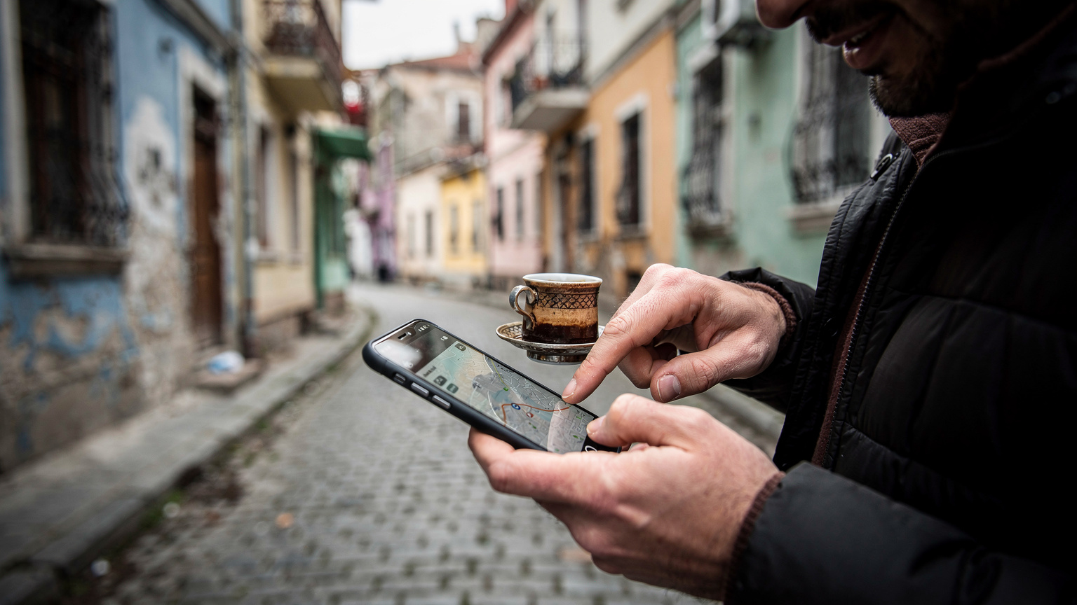 İstanbul'un tarihi Balat mahallesinde, dar ve Arnavut kaldırımlı bir sokakta yürüyen bir gezginin, elinde tuttuğu Türk kahvesi bardağıyla birlikte telefonundaki haritayı dikkatle incelediği anın yakın çekimi. Parmağı harita üzerinde bir noktayı işaret ediyor.