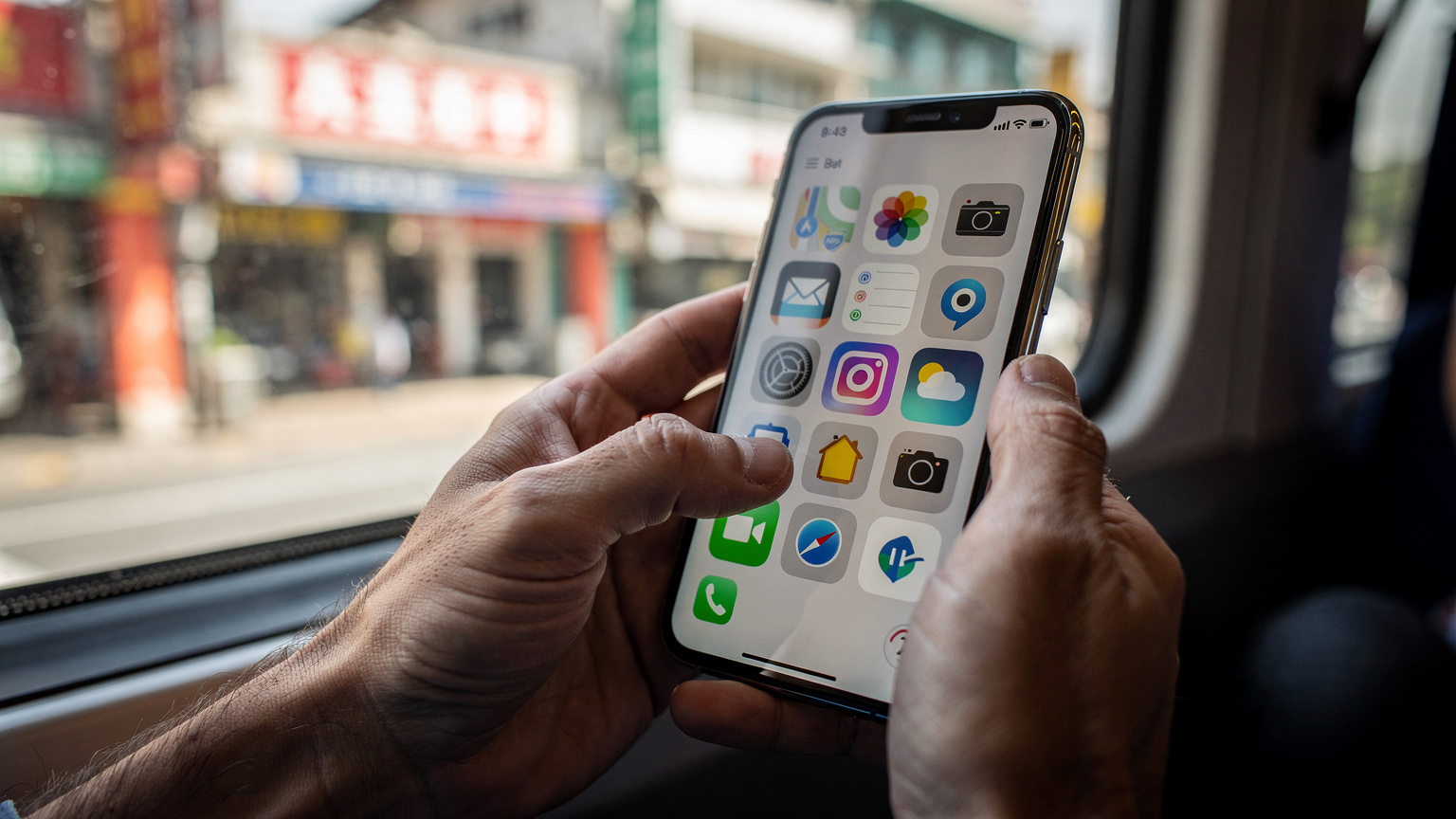 A close-up of a traveler's hands holding a smartphone, with their thumb about to tap an icon, against a blurred background of a train window looking out at a city street.