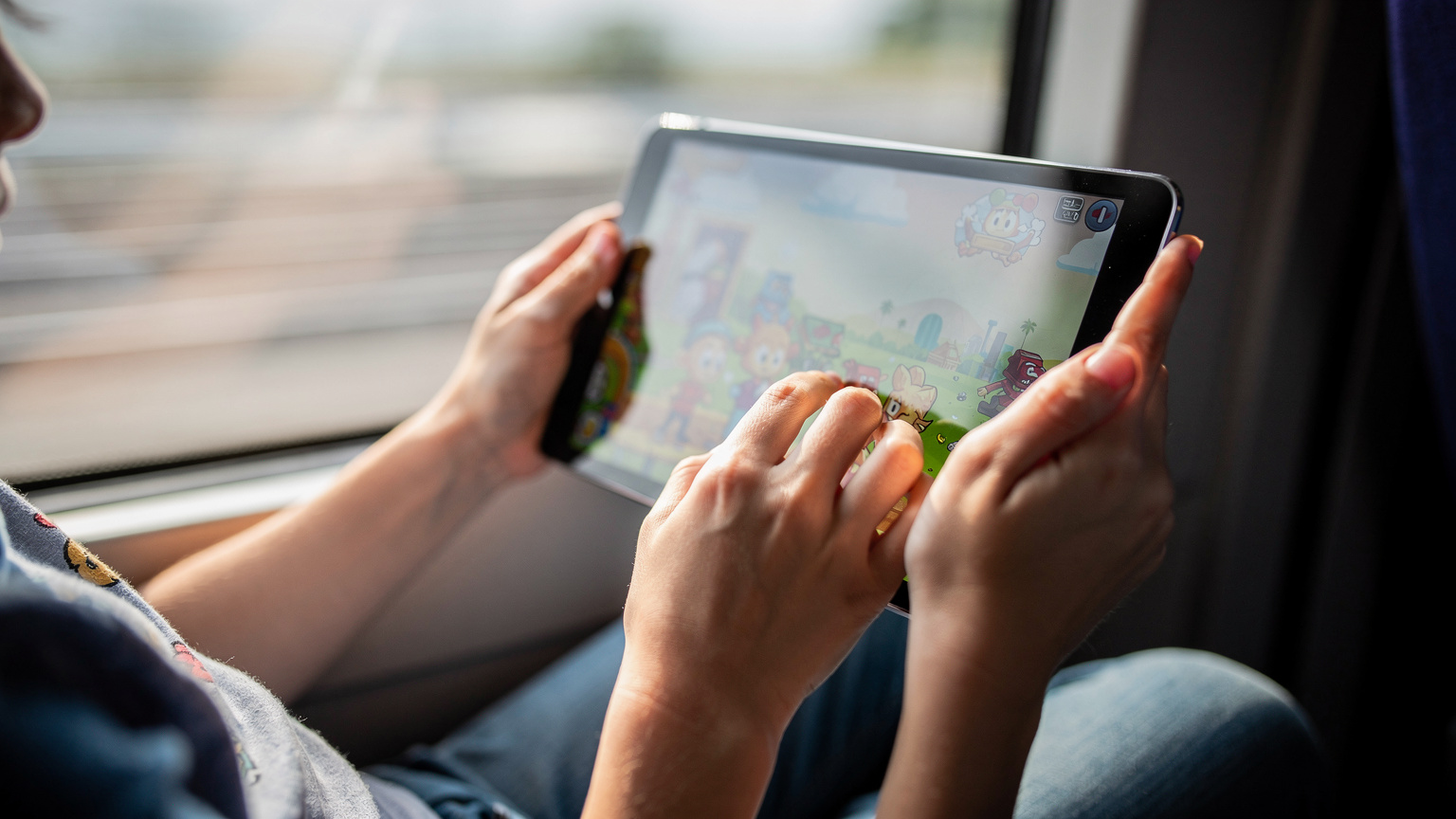 A child's hands holding an iPad, displaying a vibrant, colorful game, with a blurred background suggesting a moving train or car.