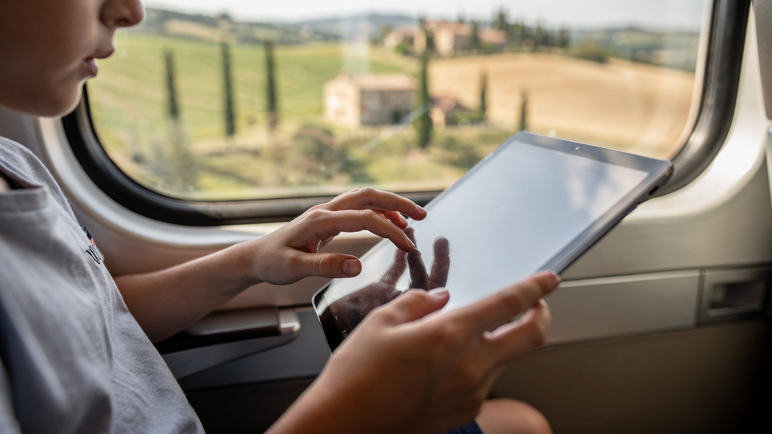 A child's hands interacting with a tablet screen while sitting on a train in Italy, with blurred Tuscan countryside visible through the window.