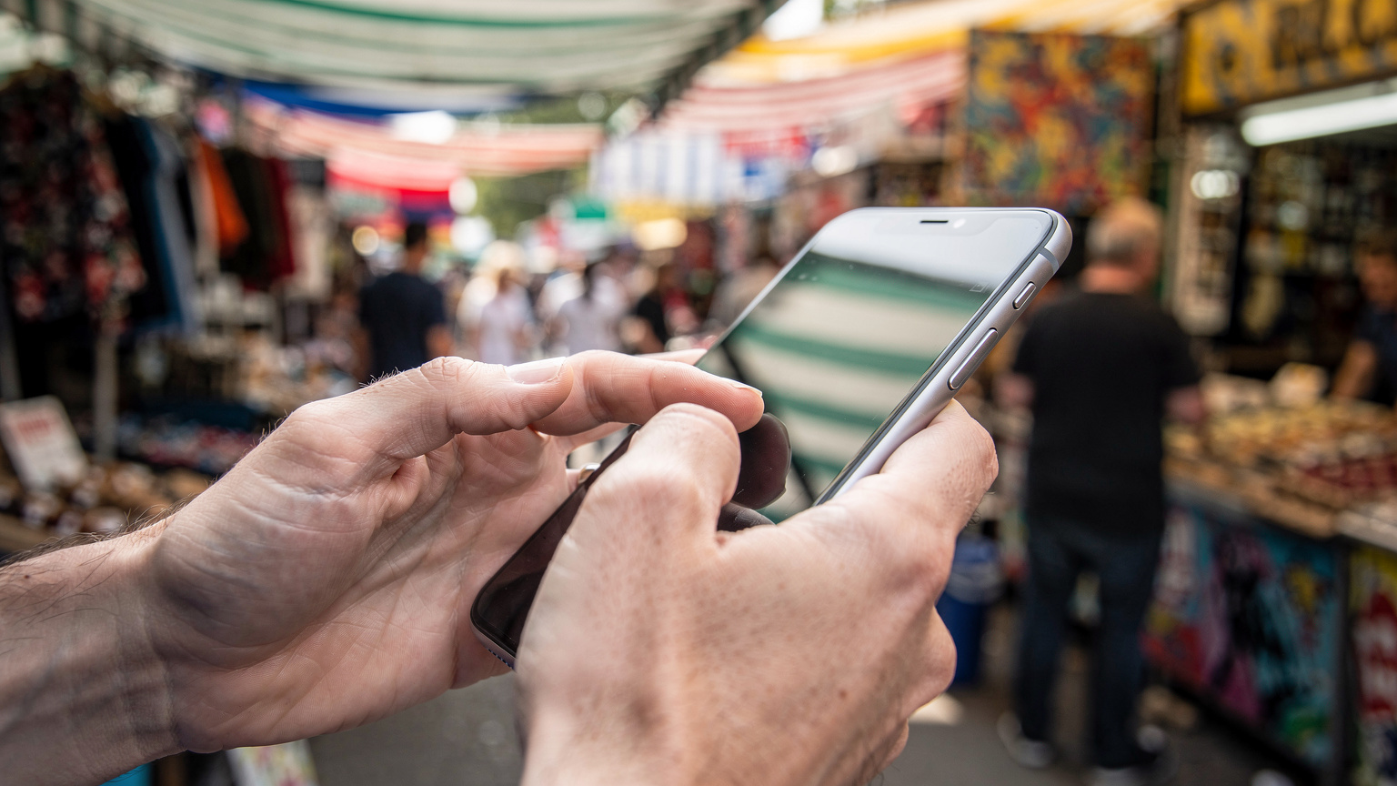 Nahaufnahme von Händen, die ein Smartphone halten, während im Hintergrund verschwommen ein Straßenmarkt in Camden Town mit vielen Menschen und bunten Ständen zu sehen ist.