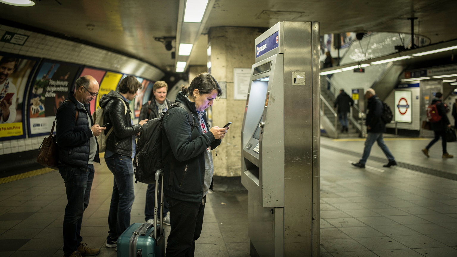 Ein Reisender versucht an einem belebten Ticketautomaten in der U-Bahn-Station Piccadilly Circus in London, sein Smartphone zu entsperren, während sich eine kleine Schlange hinter ihm bildet.