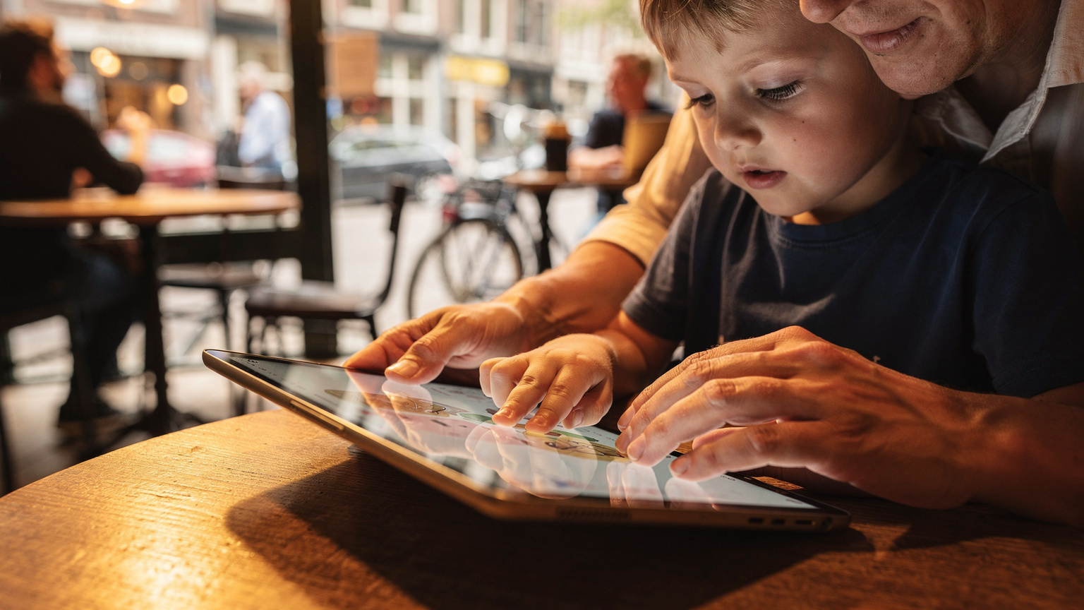 A parent's hands gently guiding a child's finger on a tablet screen, showing an educational app or offline map, set in a cozy cafe in Amsterdam, Netherlands. No screen content is legible, focusing on the interaction.