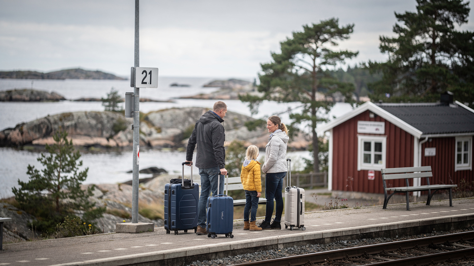 Familj med resväskor på en tågperrong i Göteborgs skärgård, klippor i bakgrunden, soft overcast ljus