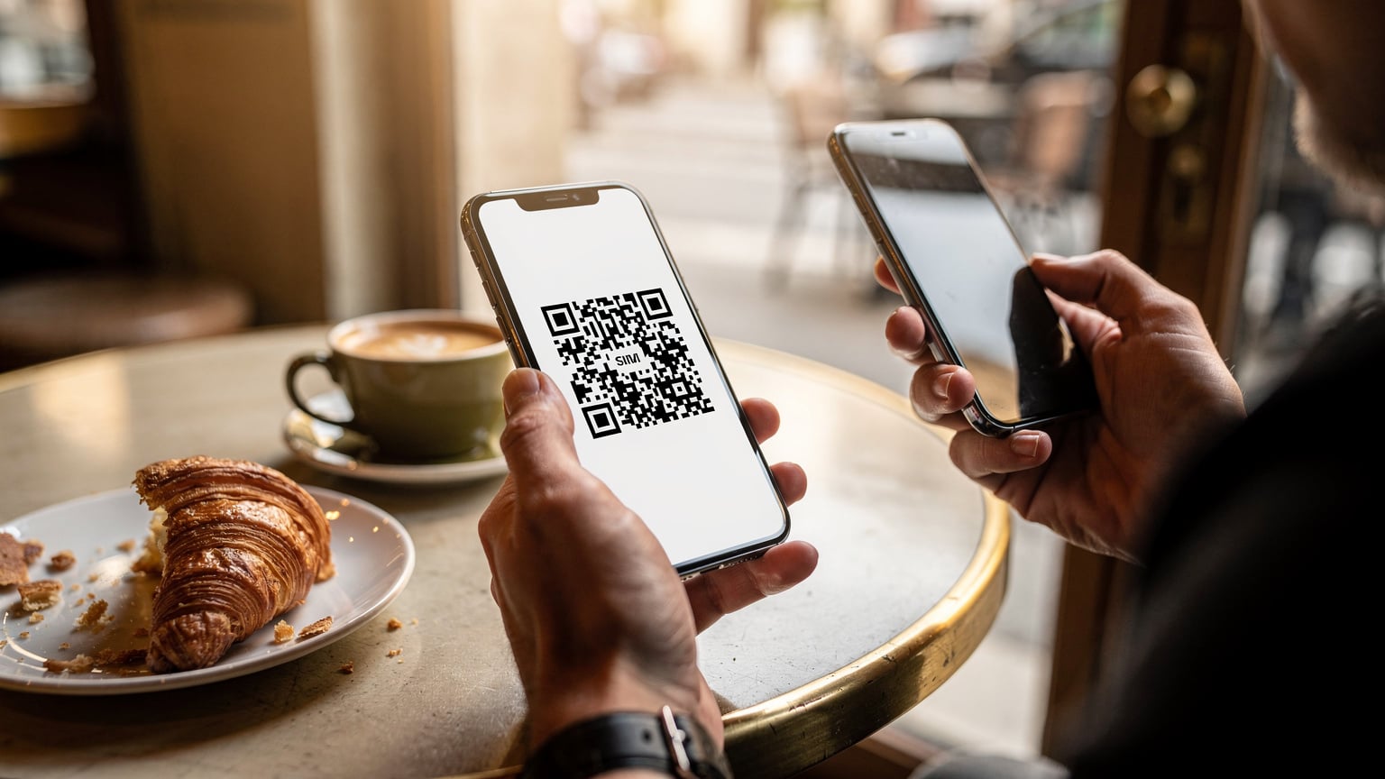 A traveler in a cafe in Paris, scanning an eSIM QR code on a smartphone with a croissant and coffee on the table.