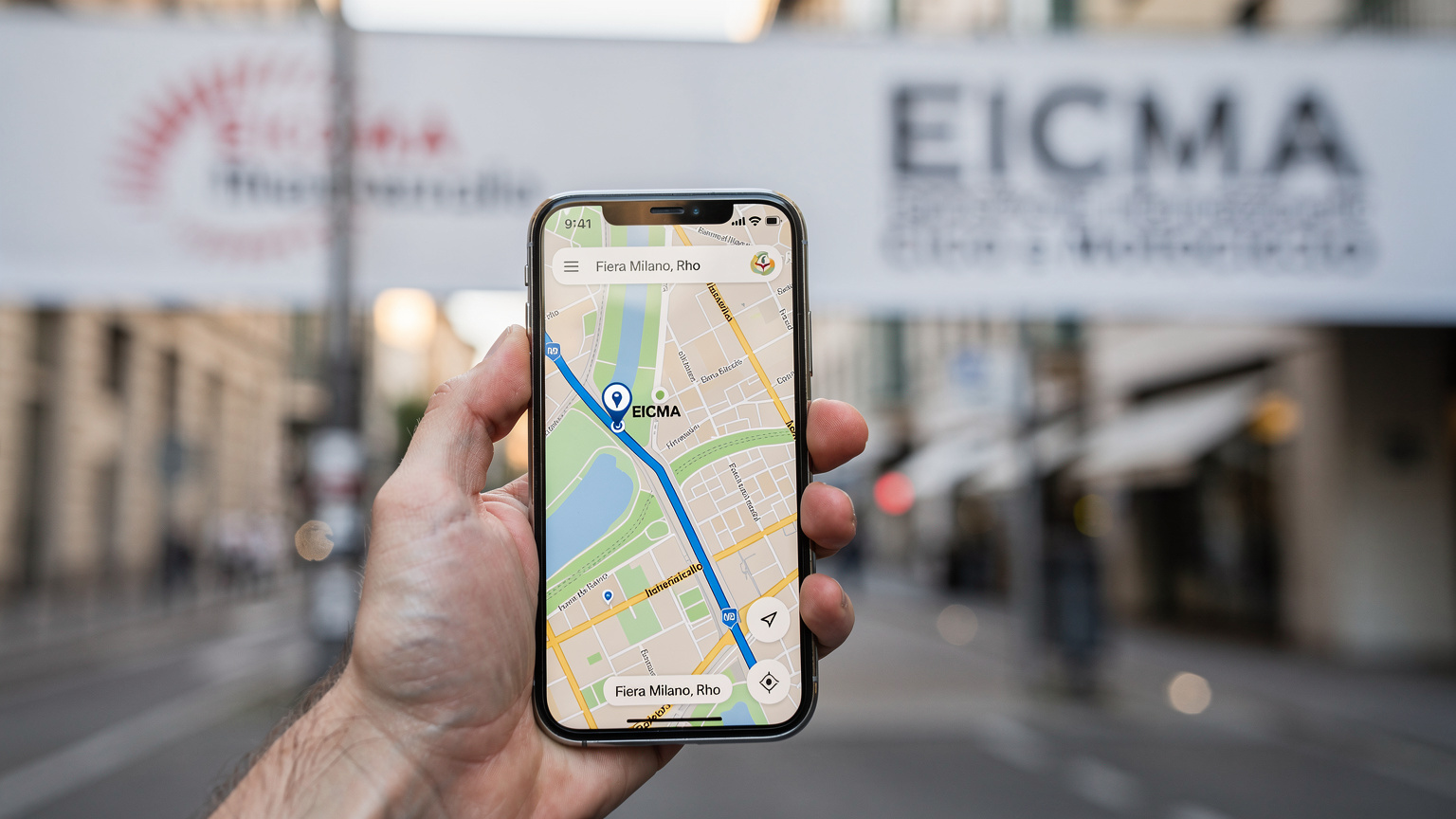 En person som navigerer på telefonen sin med et kart over Milano, med EICMA-logoen i bakgrunnen.