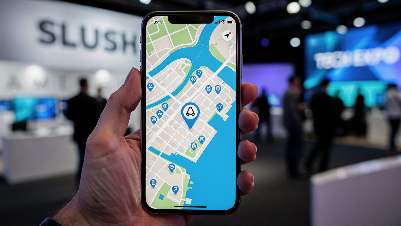 En hand håller en smartphone som visar en karta över Helsingfors, med Slush-logotypen synlig i bakgrunden, symboliserande enkel navigering och uppkoppling.