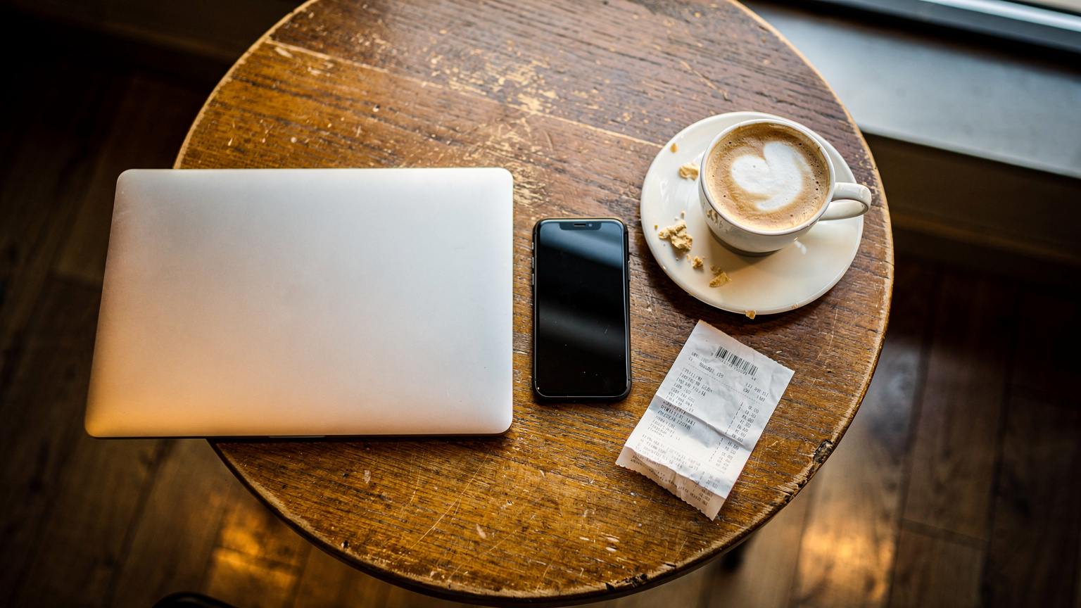 A laptop and smartphone set up on a wooden cafe table next to a flat white coffee.